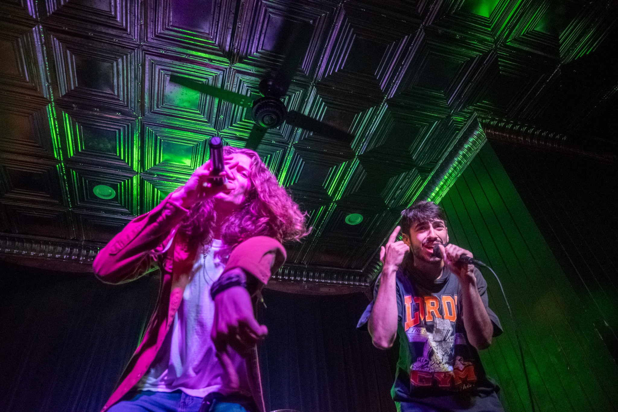 Two performers on stage under green and purple lighting, one singing into a microphone, the other gesturing with hand near ear.