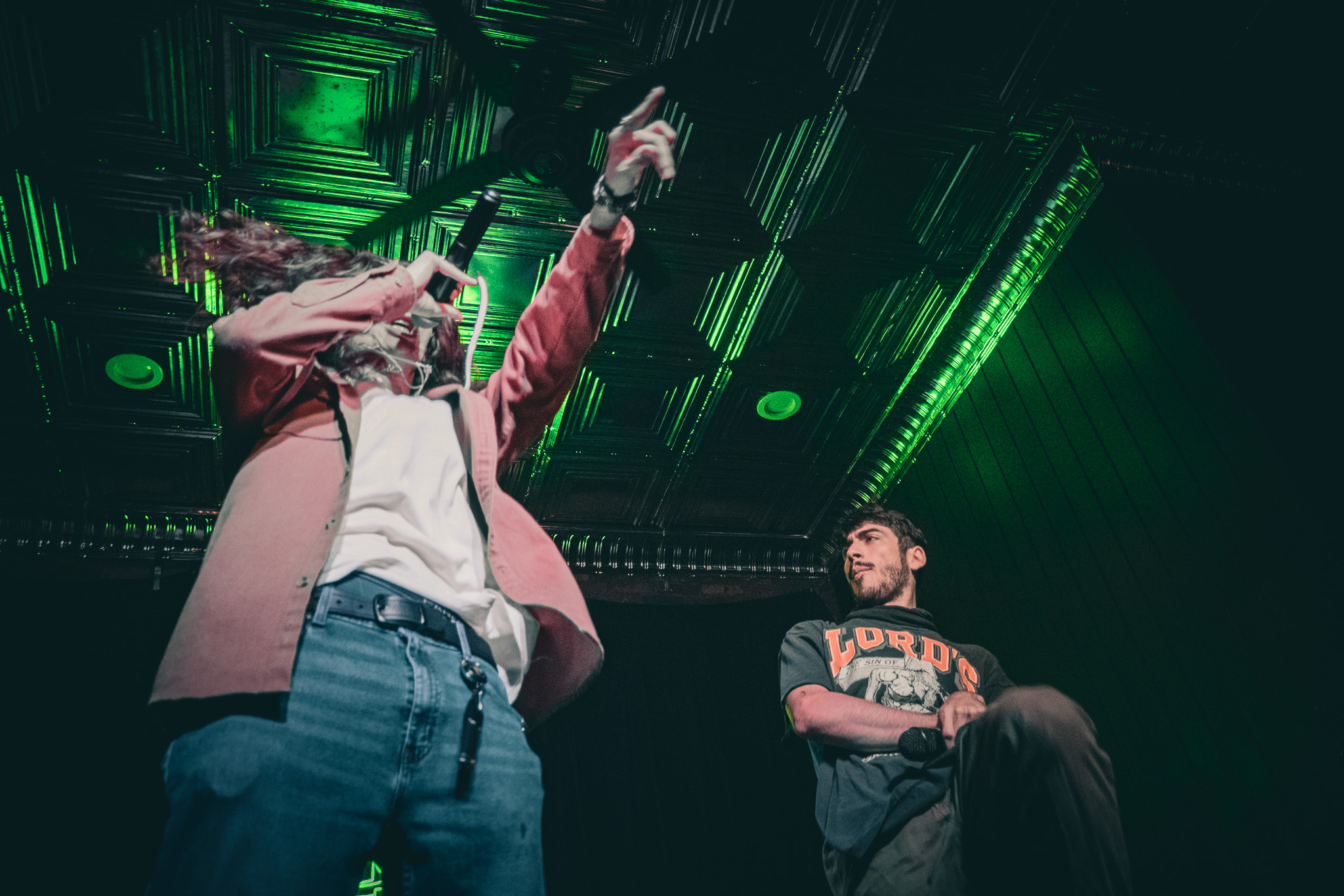 Two people in a dark room with green lighting, one pointing upward, the other standing with arms crossed.