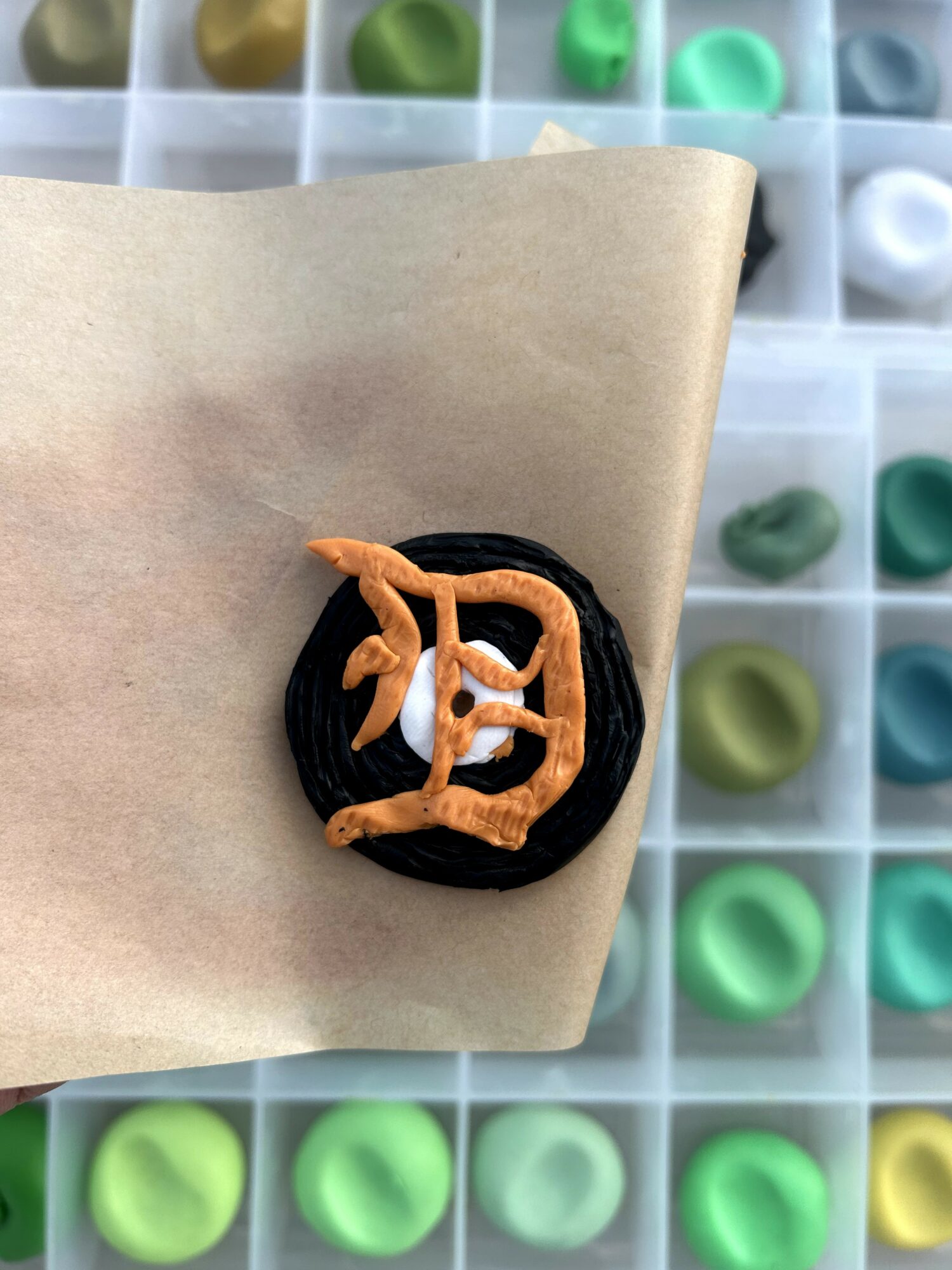 Round black object with orange and white design on beige paper, background of colorful small containers.