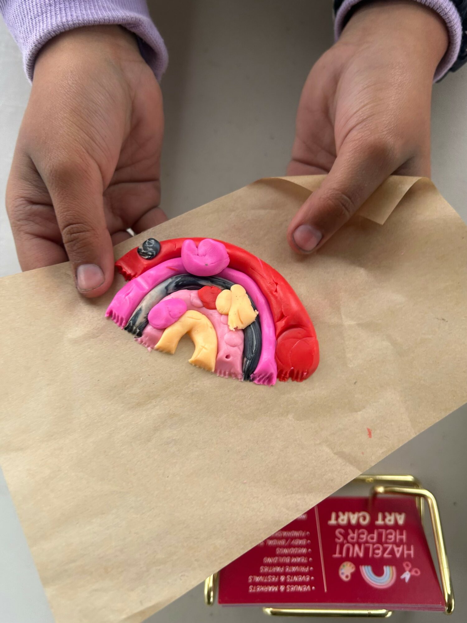 Child's hands create a rainbow with colorful clay on paper, with a red box nearby, in a craft activity.
