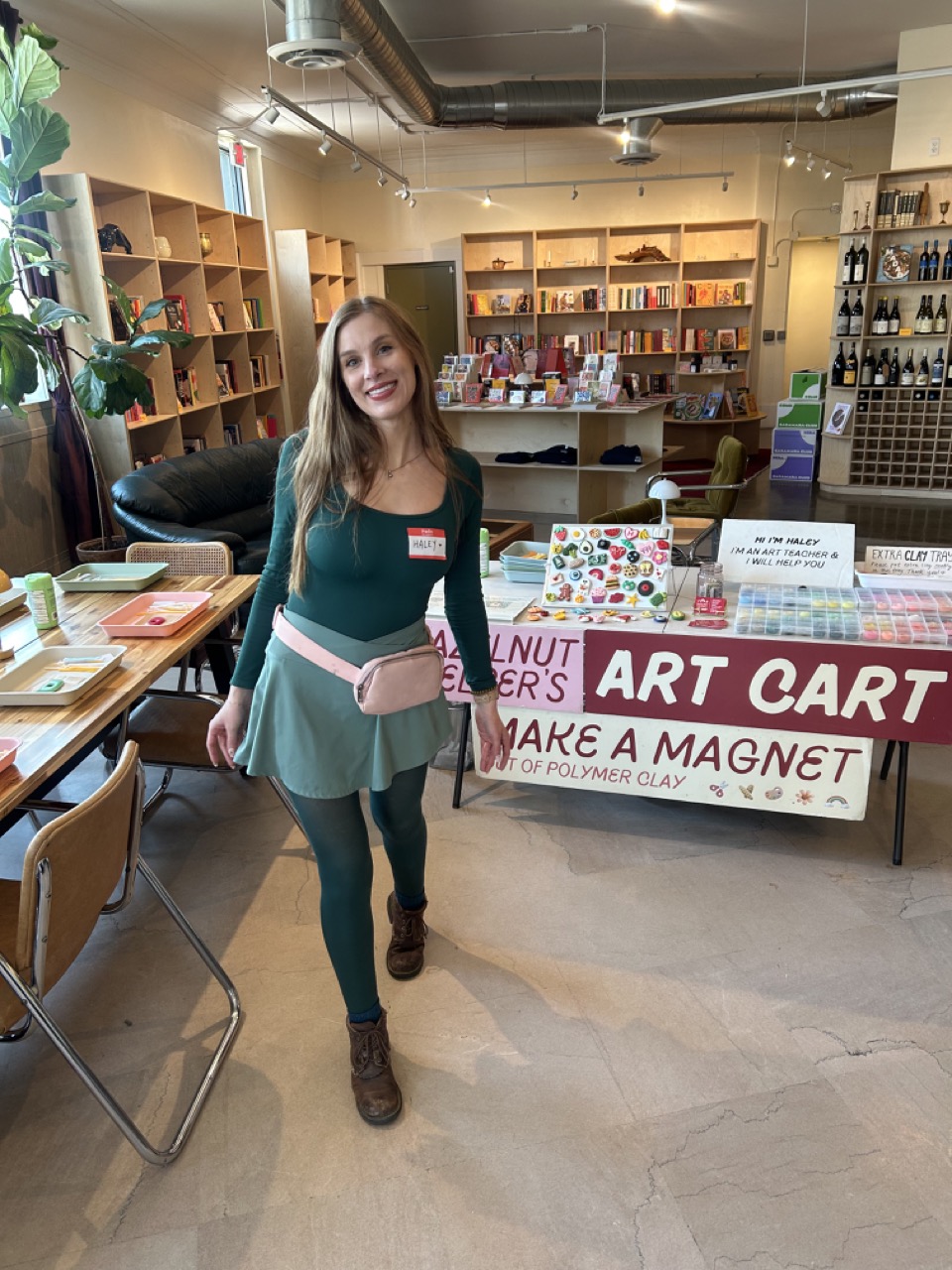 Young woman with long hair smiling, standing in a bookstore or craft store, holding a pink bag, surrounded by shelves and tables.