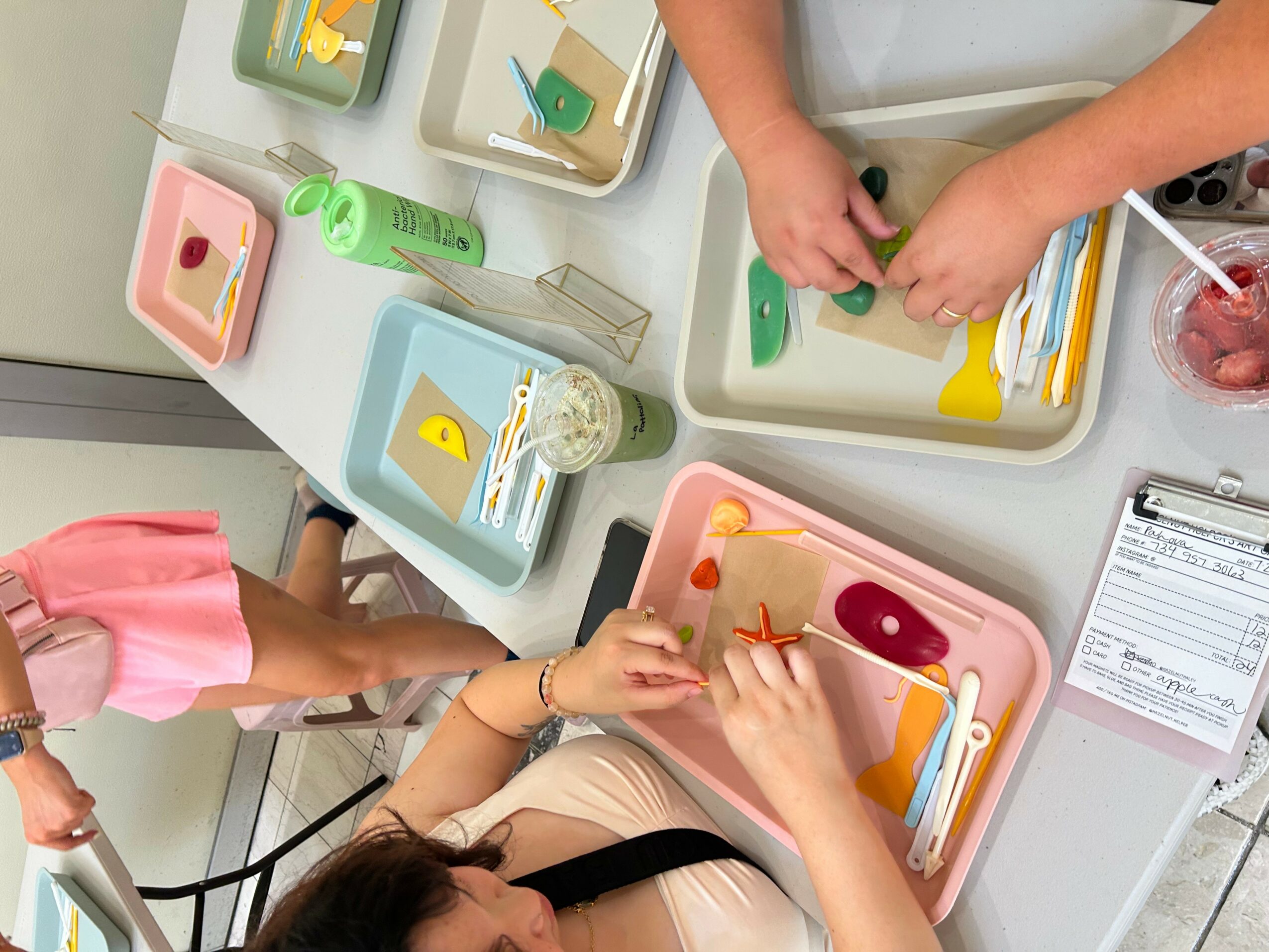 Hands preparing food on a tray with various utensils and ingredients, including a person in pink and another in beige, in a kitchen.