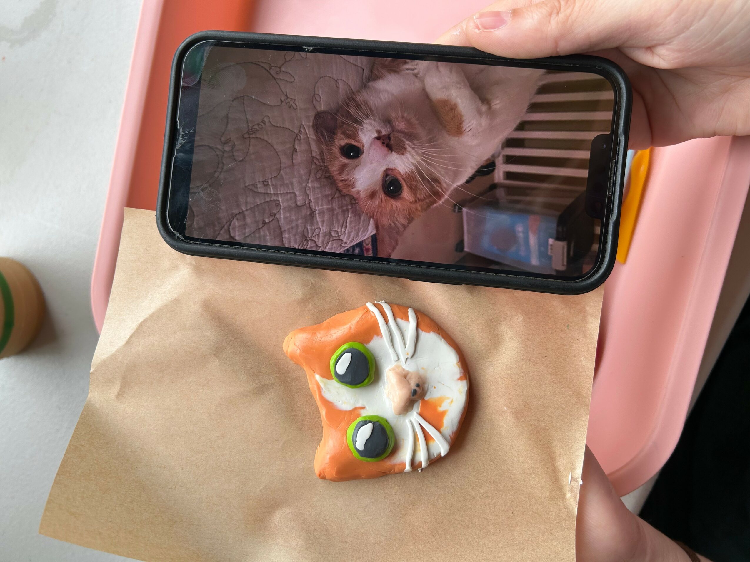 Person holding a smartphone showing a dog, with a small orange and white cat-shaped object on a paper surface below.