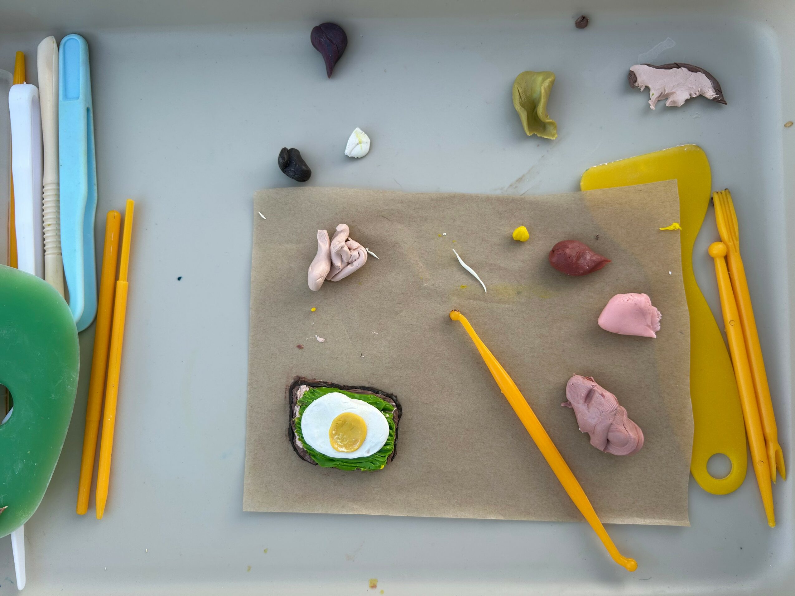 Colorful modeling clay and tools on a tray with a paper sheet, including a clay egg, animal shapes, and sculpting tools.