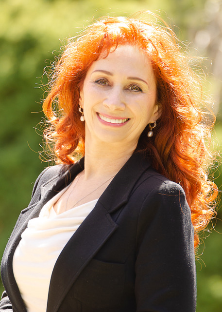 Woman with curly red hair smiling outdoors, wearing a black blazer and white top, sunlight and greenery in background.