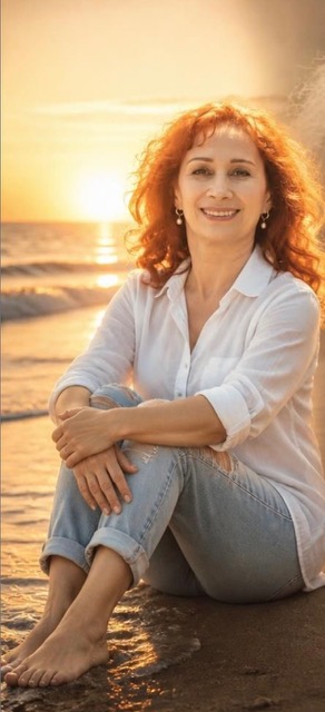 Woman with curly red hair sitting on the beach at sunset, smiling, wearing a white shirt and jeans.