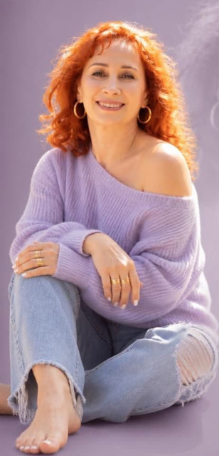 Woman with curly red hair sitting barefoot, wearing a purple sweater and ripped jeans, smiling outdoors.