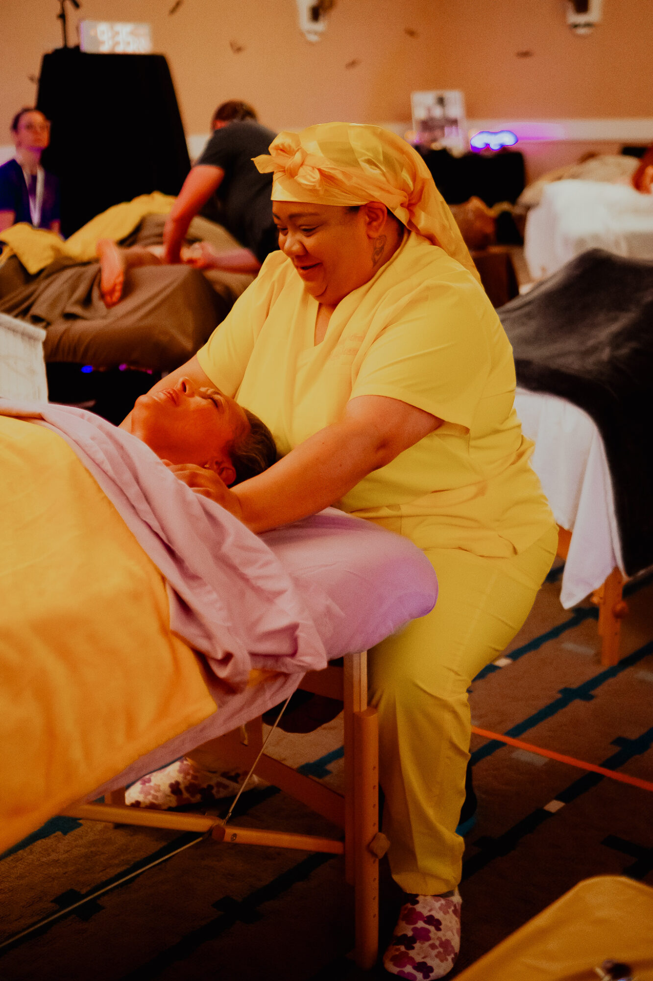 Healthcare worker in yellow scrubs and head covering assisting a patient on a bed in a busy medical setting.