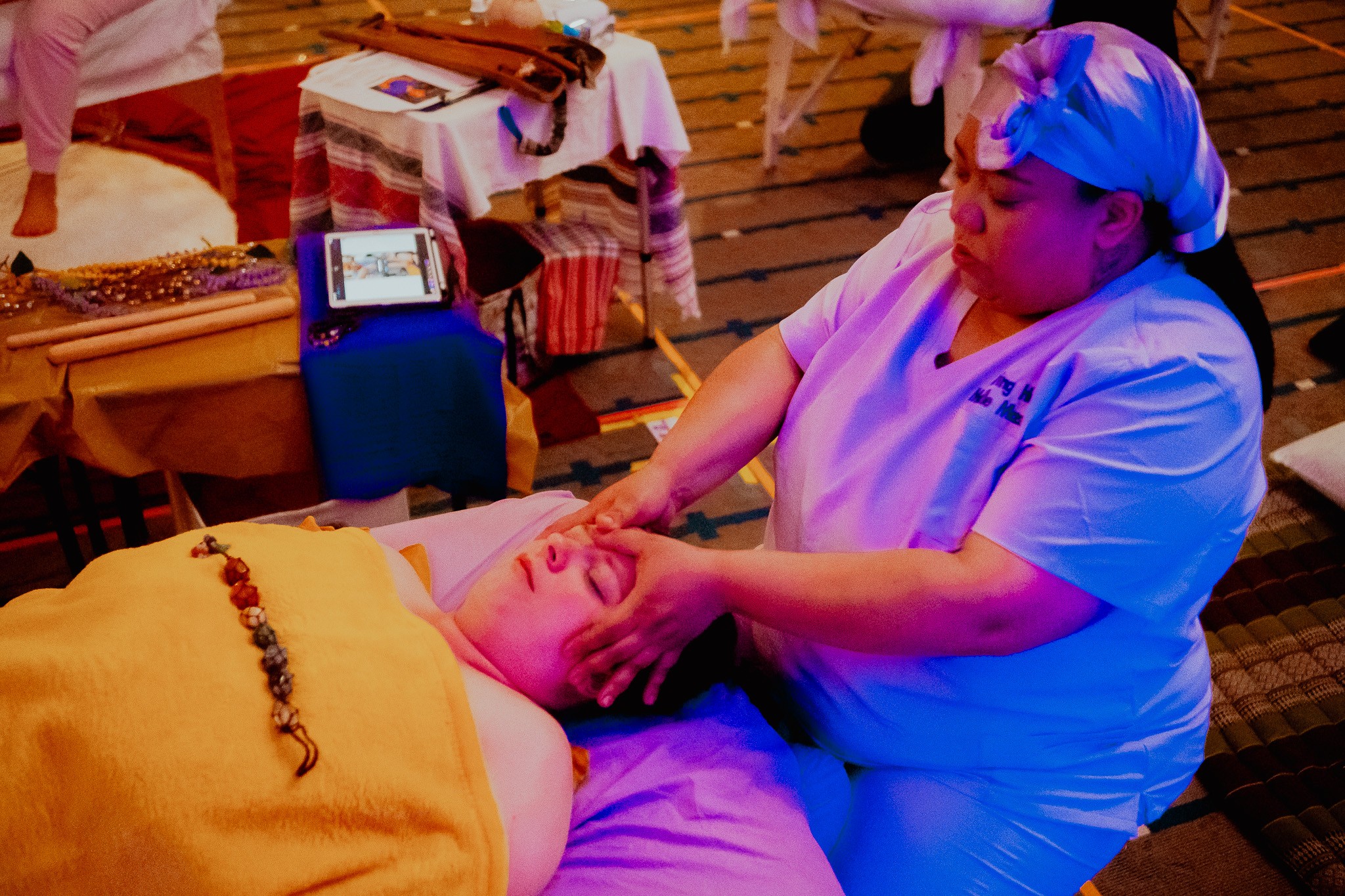 Person receiving a massage from a massage therapist in a room with wooden floors and tables, warm lighting.