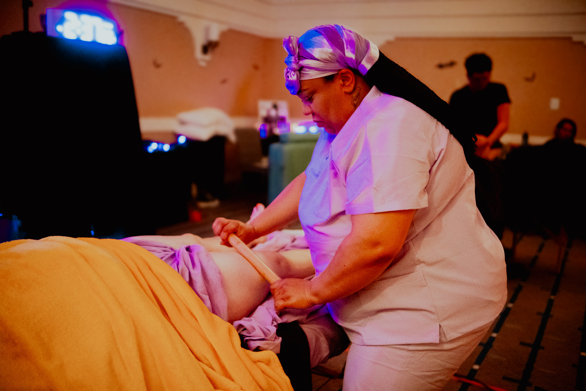 Nurse performing a medical procedure on a patient lying on a bed in a room with other people in the background.