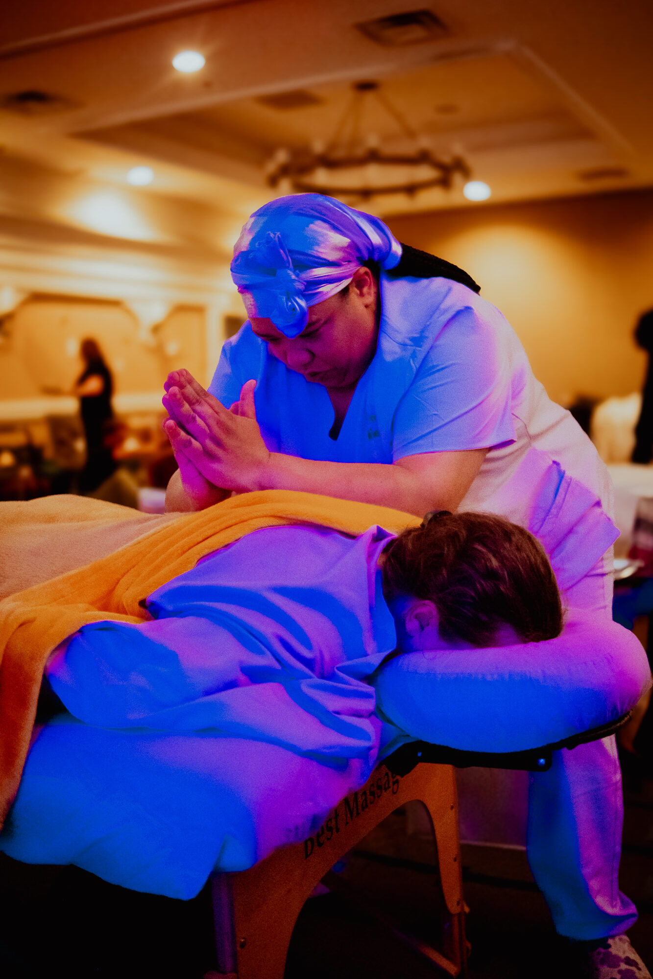 Healthcare worker in blue scrubs and cap performs a procedure on a patient lying face down on a massage table in a dimly lit room.