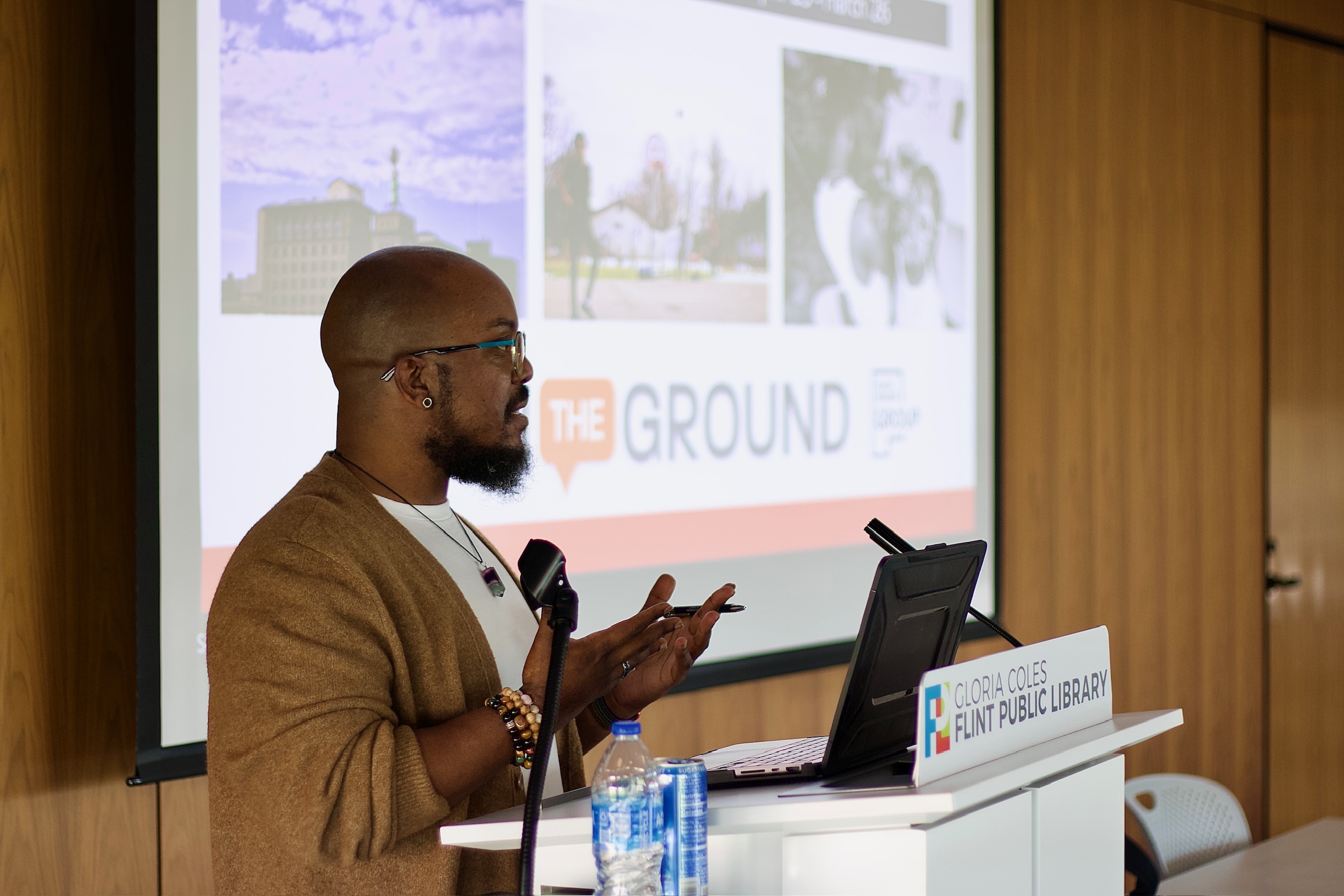 Man giving a presentation in front of a screen, with a laptop and water bottles on the table, in a conference room.