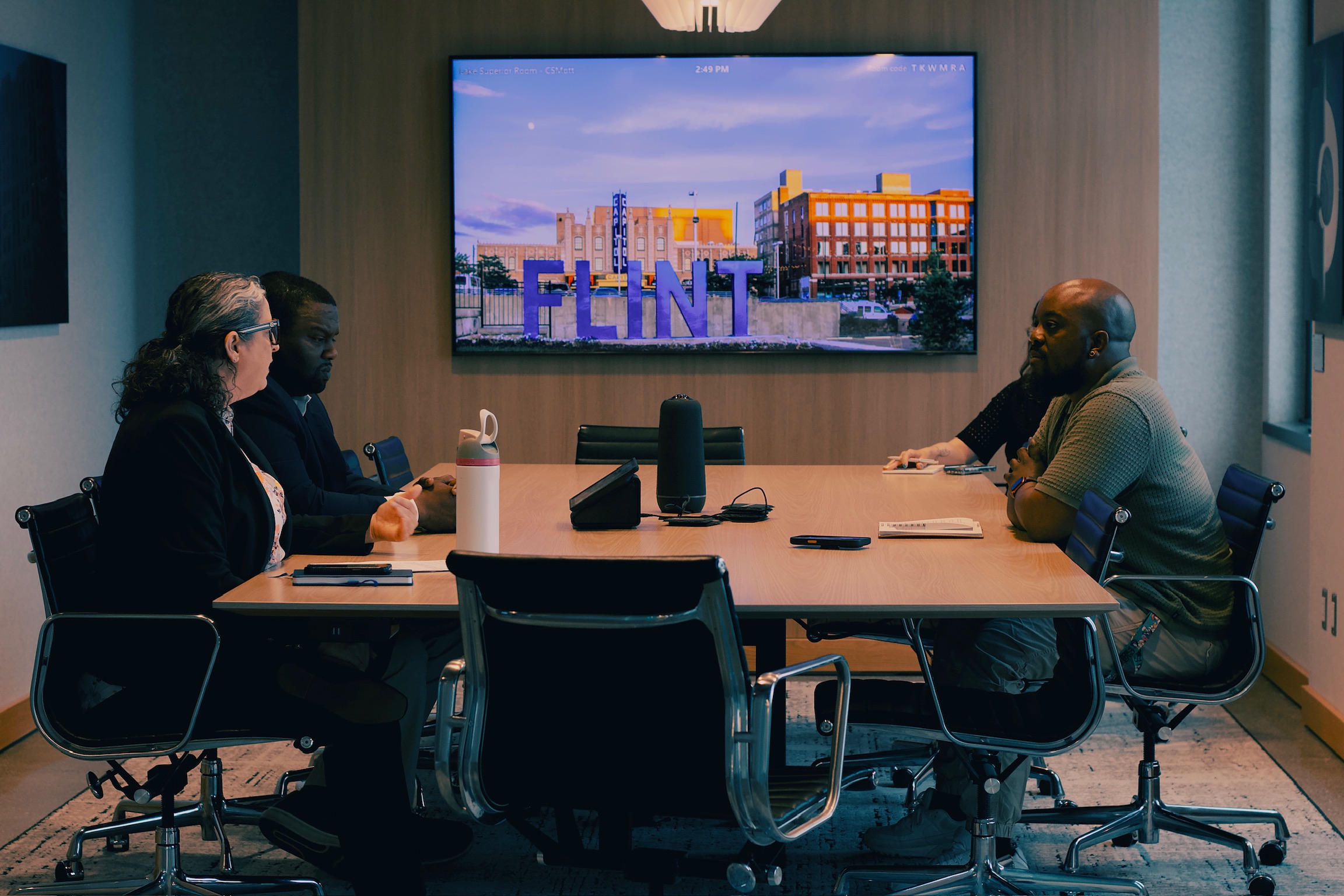 Four people sit around a conference table watching a large screen displaying the word 'FLINT' and a cityscape.