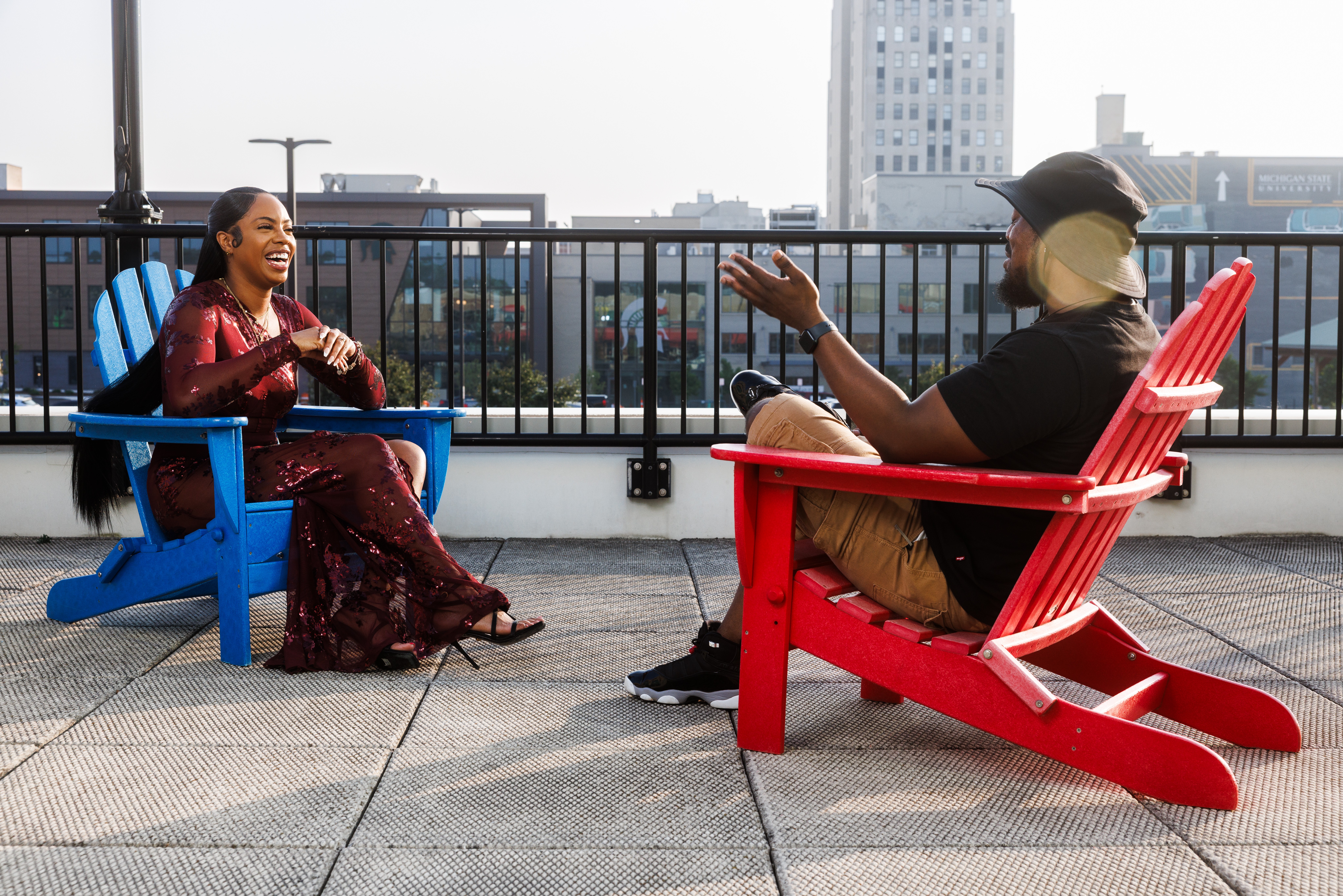 Two people sitting on colorful chairs on a rooftop, engaging in conversation with city buildings in the background.