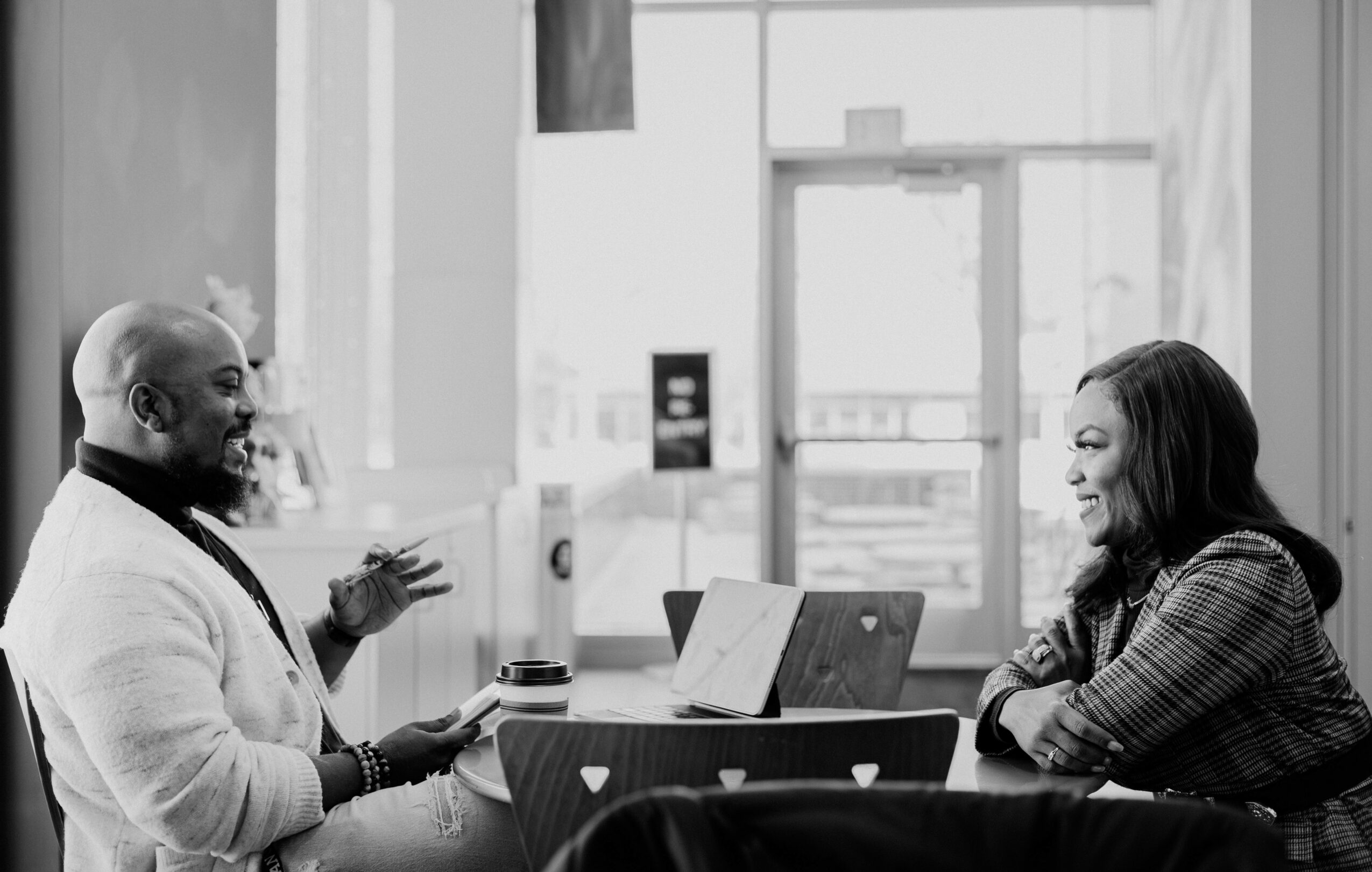 Two people sitting at a table in a bright room, engaging in conversation with laptops and a cup on the table.