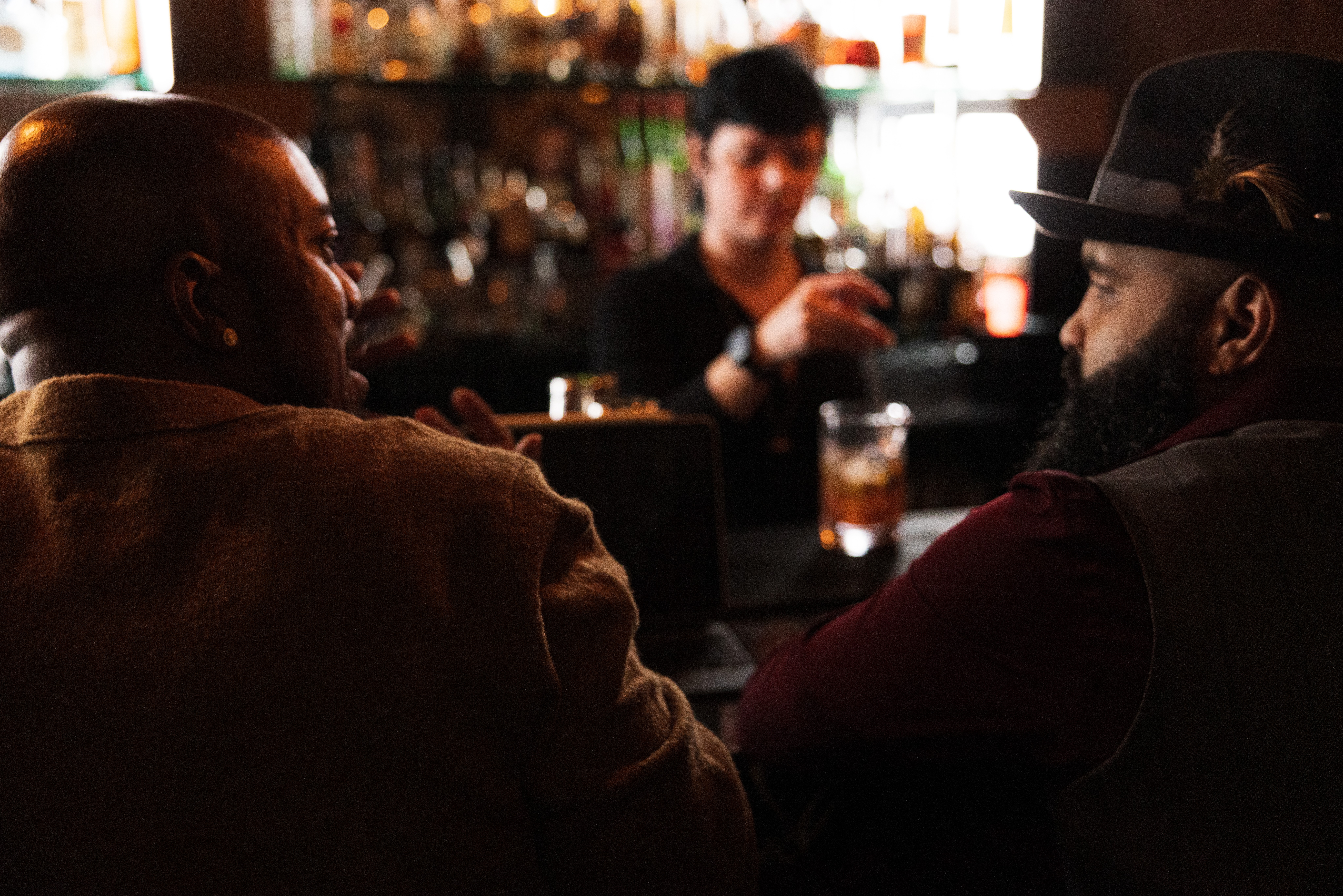 Two men sit at a bar, talking, with a bartender in the background holding a drink, in a dimly lit setting.