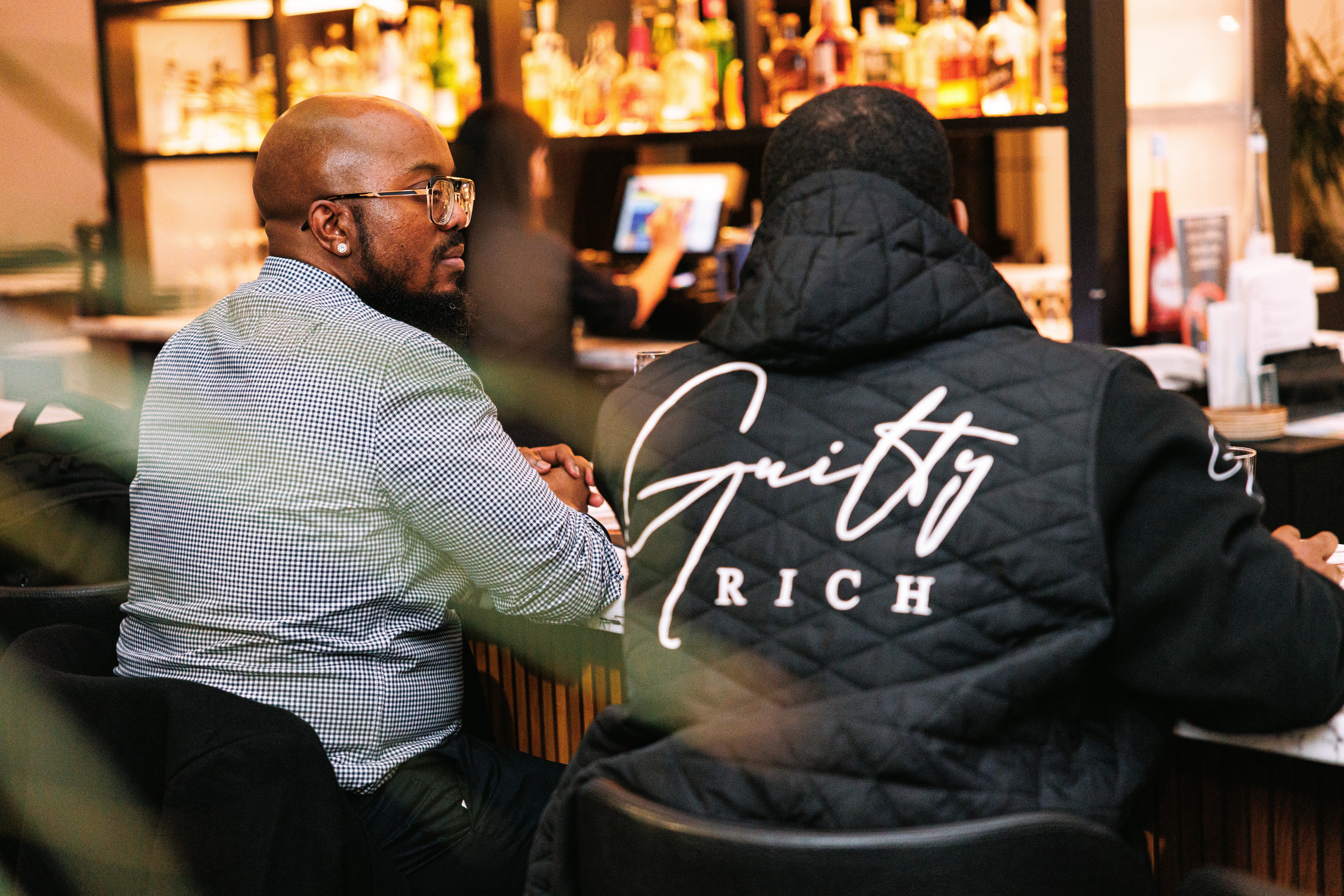 Two men sit at a bar, one wearing a gray shirt and glasses, the other in a black hoodie with 'Gritty Rich' on back, in a well-lit bar setting.