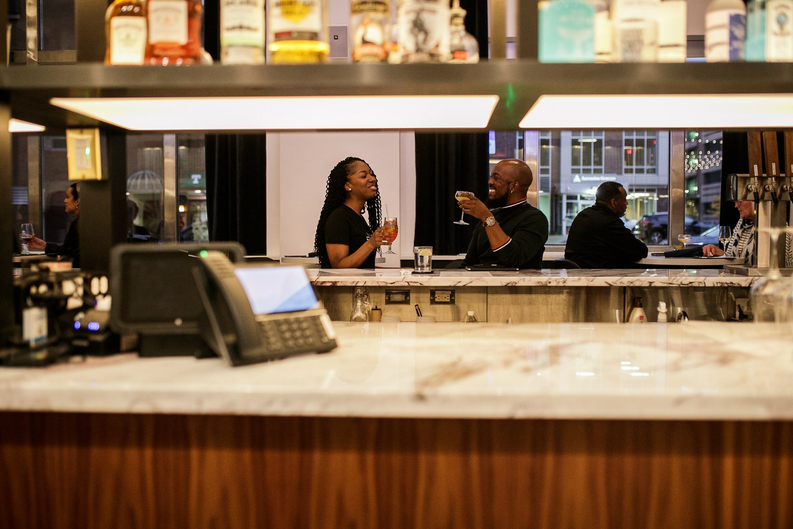 Two people sit at a bar counter, talking and drinking, with bottles displayed above and a window behind them.