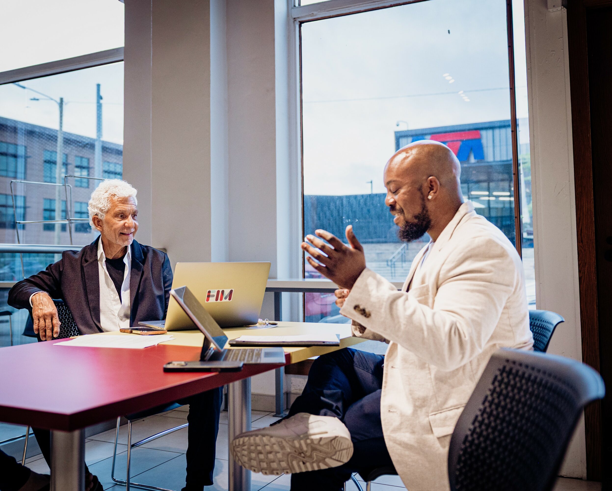 Two people in a meeting room with large windows, one elderly woman and one man in a white jacket, talking.
