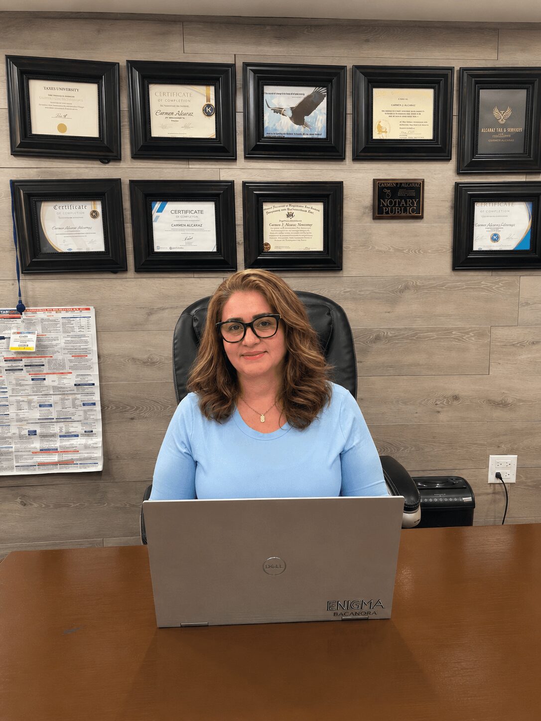 Woman with glasses sitting at a desk with a laptop, framed certificates on the wall behind her.
