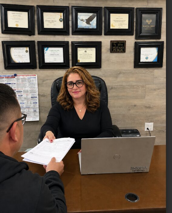 Woman with glasses and wavy hair sitting at desk with laptop, talking to a person holding papers, framed certificates on wall behind.