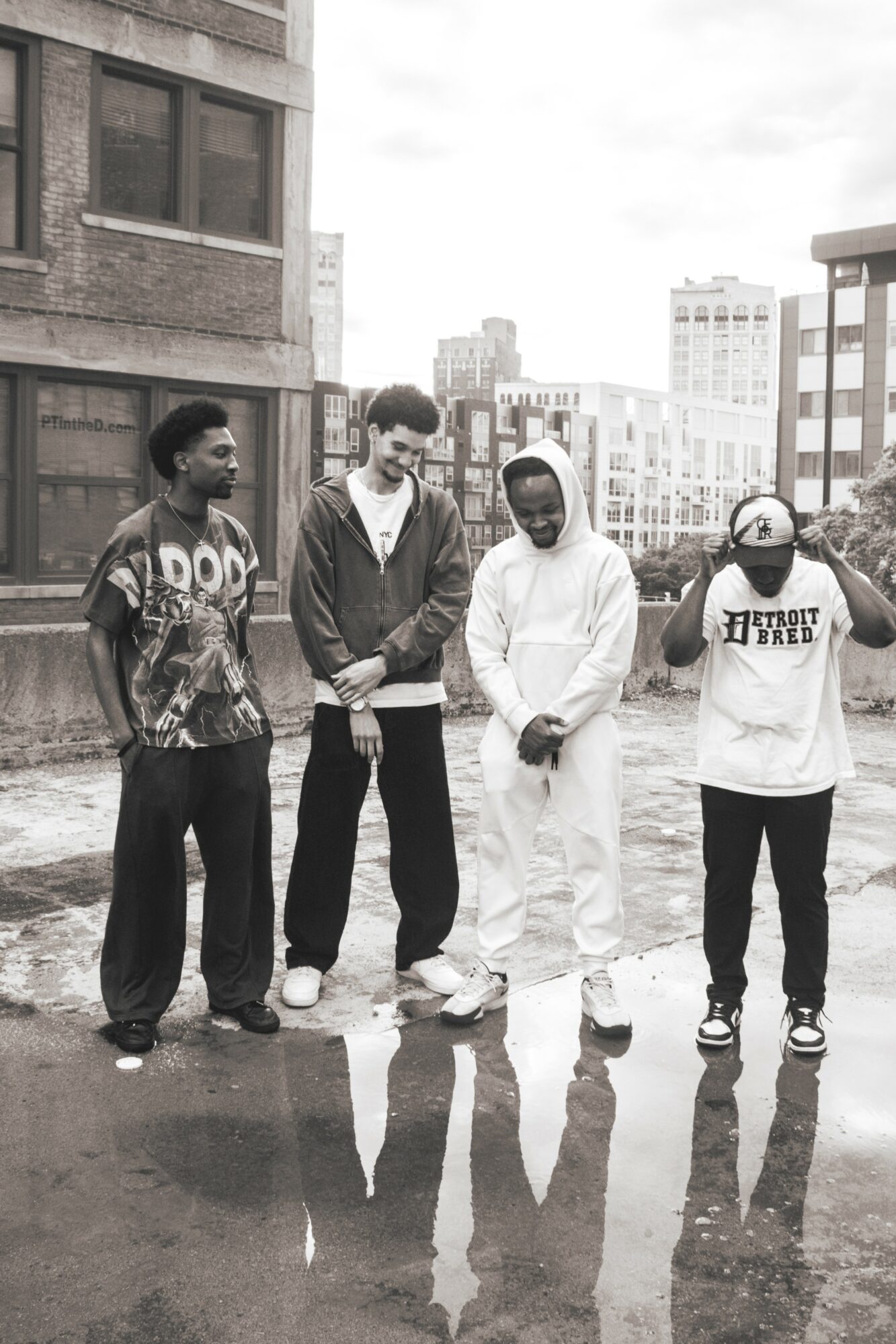 Four people stand on a rooftop with city buildings in the background, reflections visible on wet ground, black and white photo.
