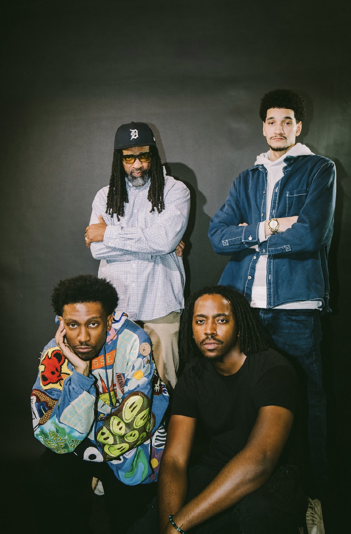 Four young men pose against a dark background, with two standing and two sitting, dressed casually.