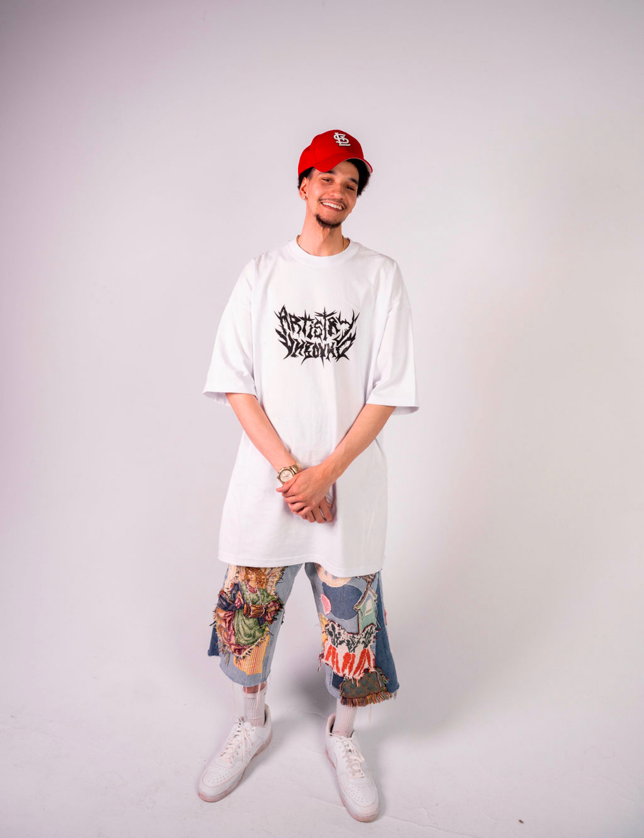 Person smiling, wearing a red cap, white oversized T-shirt, colorful patterned pants, and white sneakers, standing against a plain background.