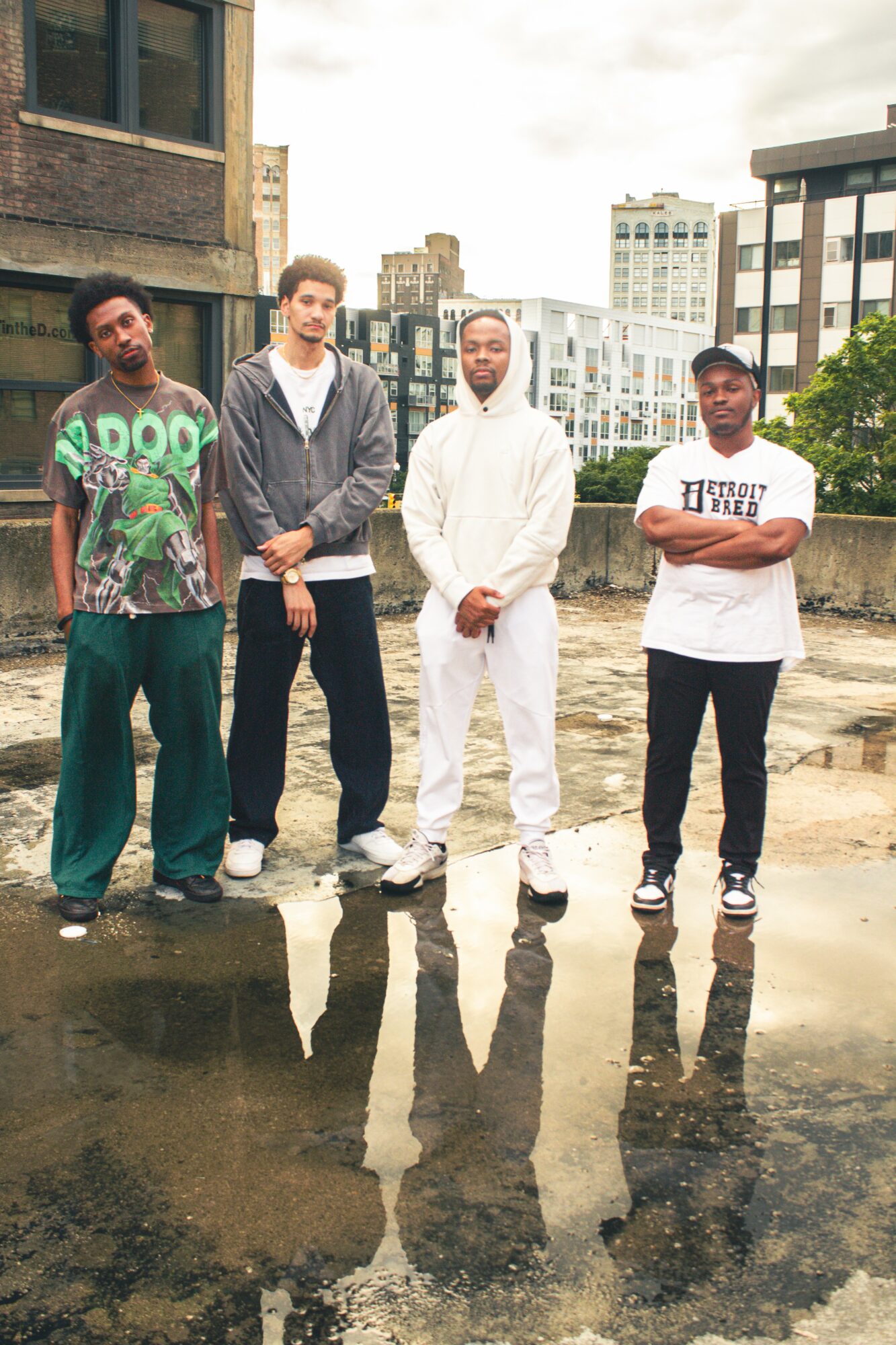 Four people standing outdoors on a rooftop with city buildings in the background, reflected in a puddle, overcast sky.