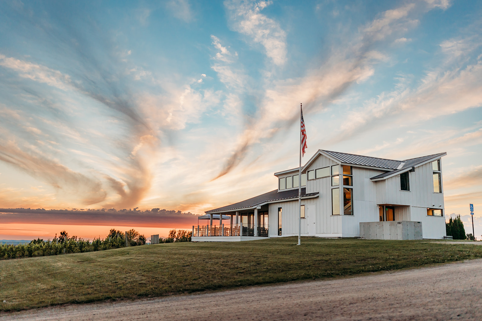 White house with porch and flagpole, overlooking water and sunset sky, grassy area in foreground.