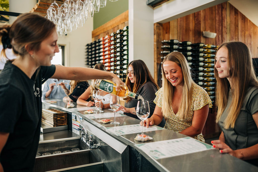 A woman pours wine into glasses at a bar with three women smiling and talking, wine bottles on shelves behind them.