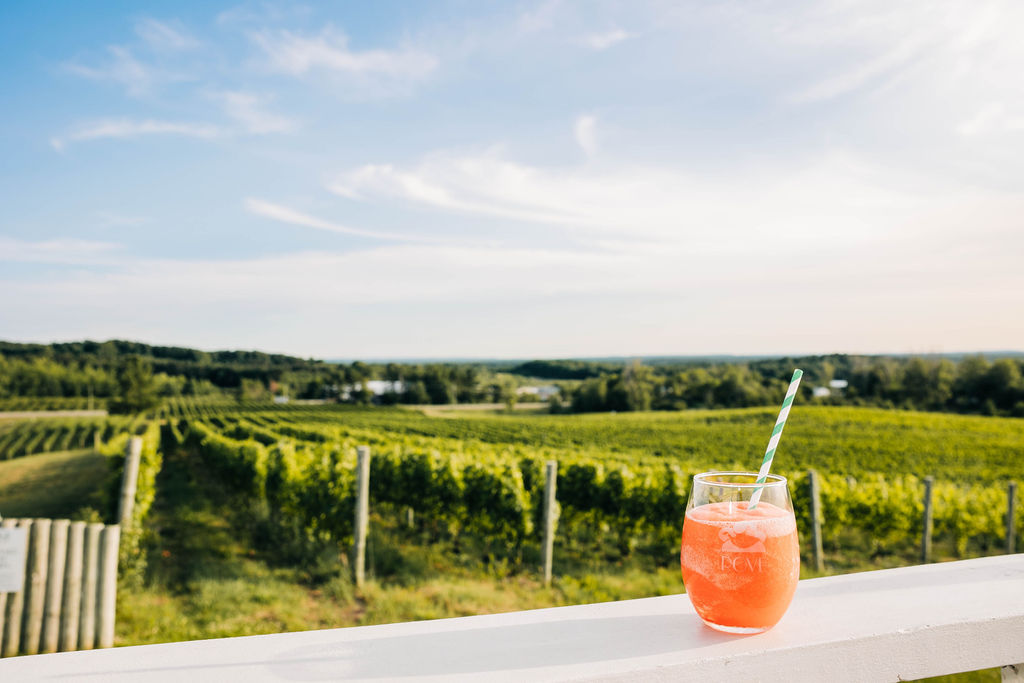 A pink drink with a straw on a white railing overlooking a vineyard landscape under a partly cloudy sky.