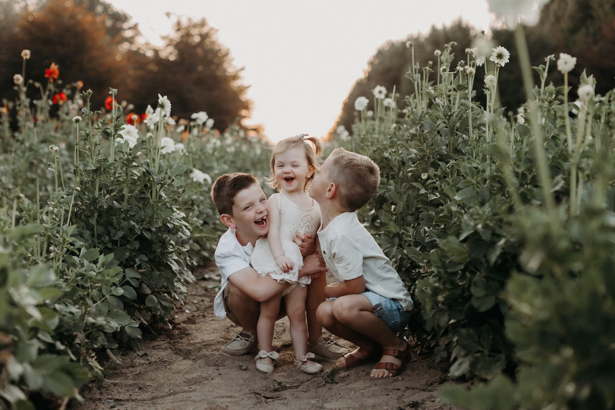 Three children laughing and playing in a garden with tall plants and flowers, sunlight in the background.