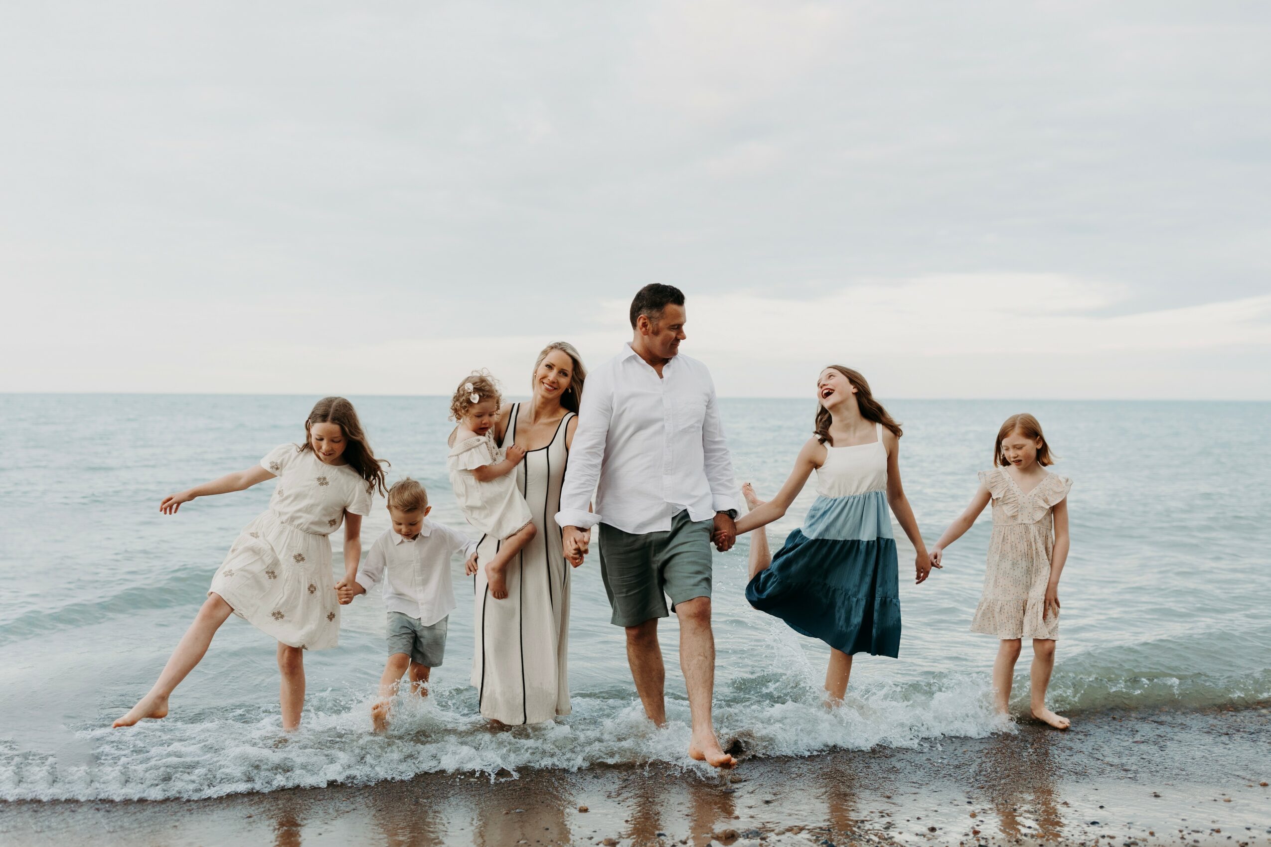 Group of six people, including children and adults, walking along the beach holding hands, with ocean and cloudy sky in background.