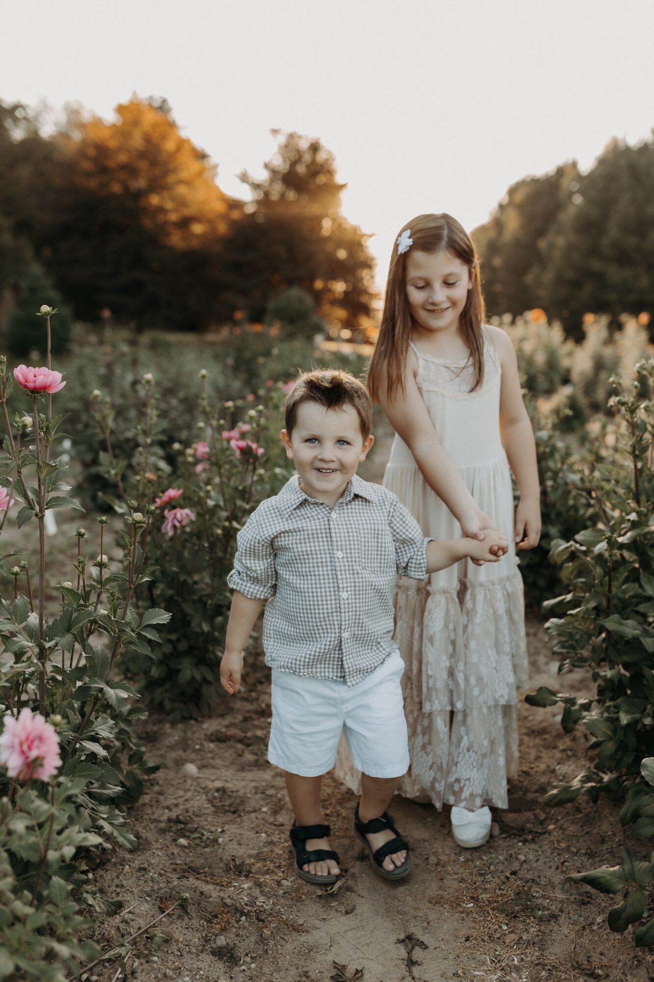 Two children, a girl and a boy, holding hands in a garden with flowers, during sunset, smiling.