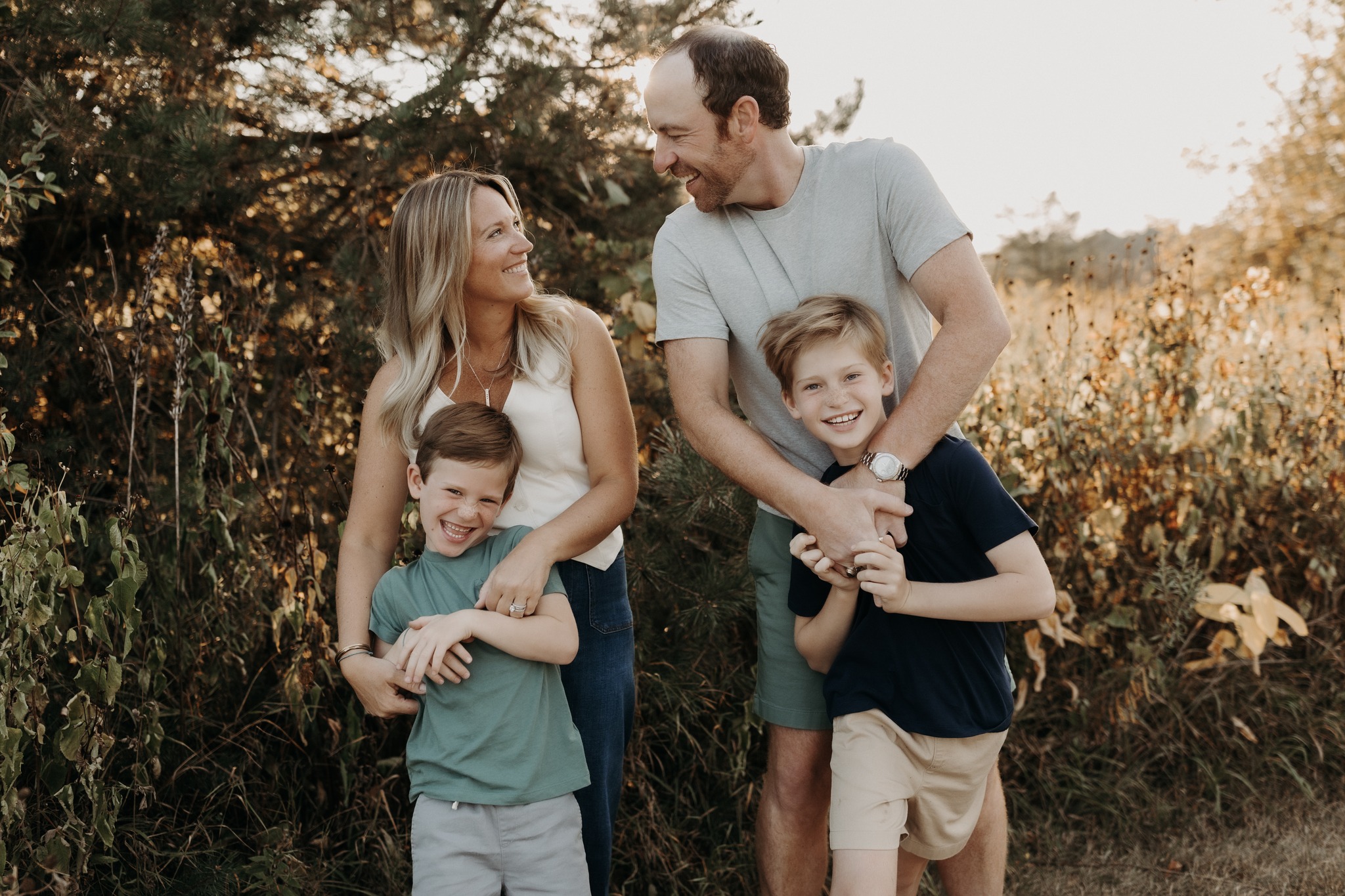 Family of four outdoors, smiling and interacting, with trees and foliage in the background.