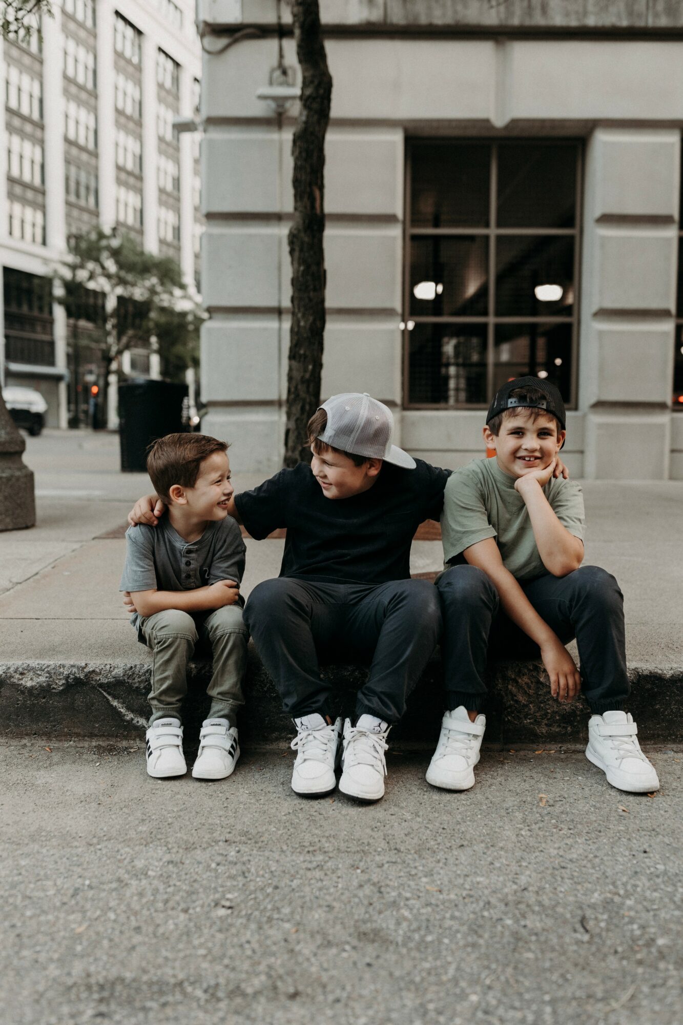 Three boys sitting on a sidewalk curb in an urban area, engaging in conversation, with buildings and a tree in the background.