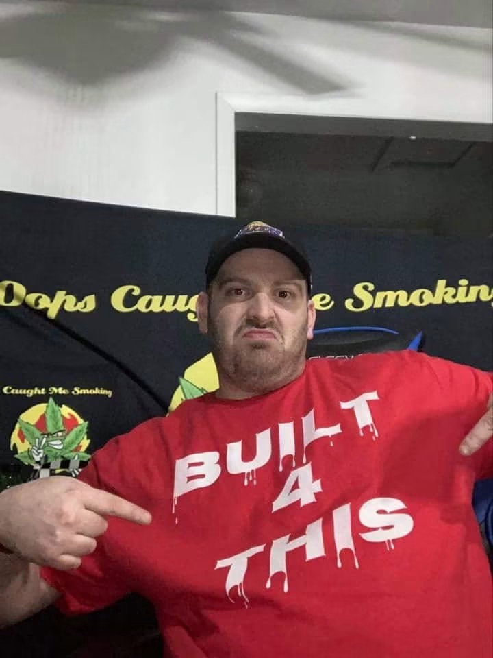 Man wearing a red shirt with white text, pointing at his shirt, in front of a black background with yellow text.
