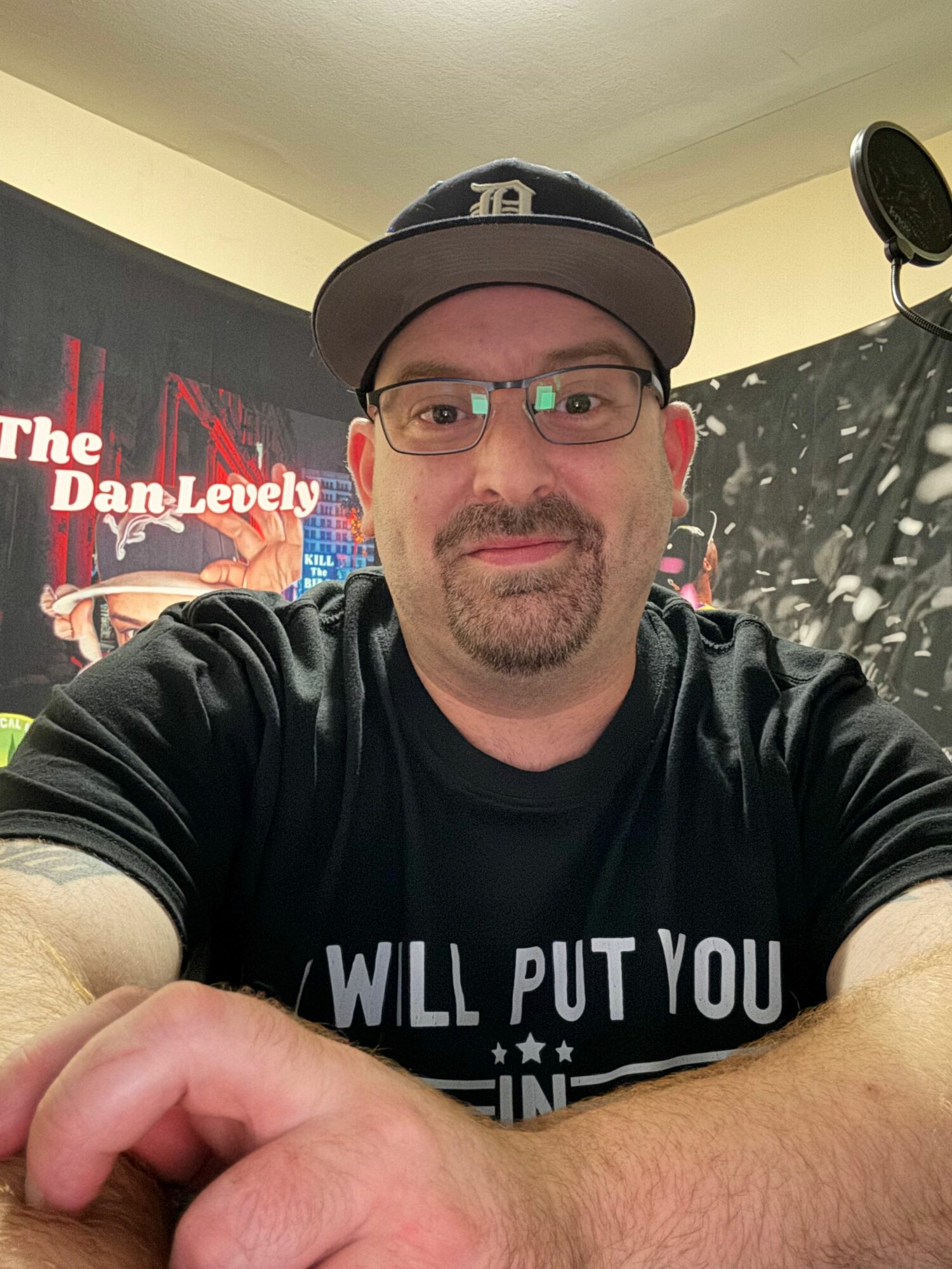 Man wearing glasses and a cap, sitting at a table with a poster in the background, looking at the camera.