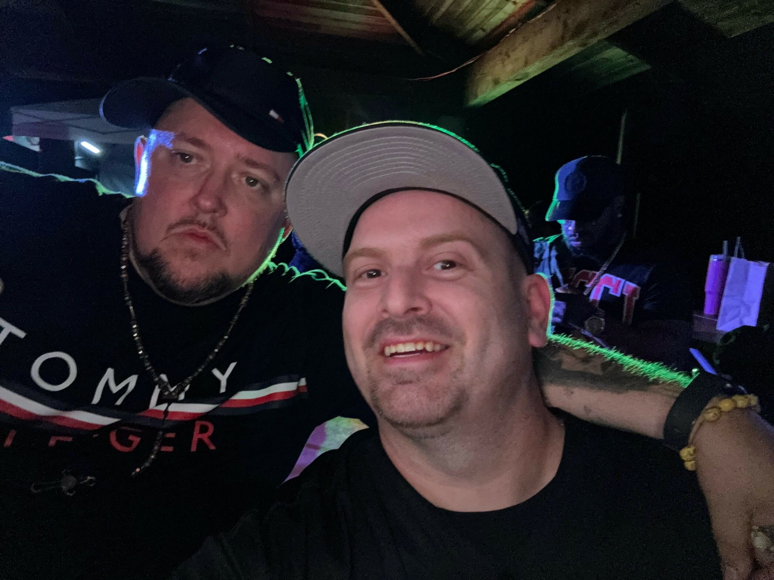 Two men posing closely, one with a serious expression and the other smiling, in a dark indoor setting with colorful lights.