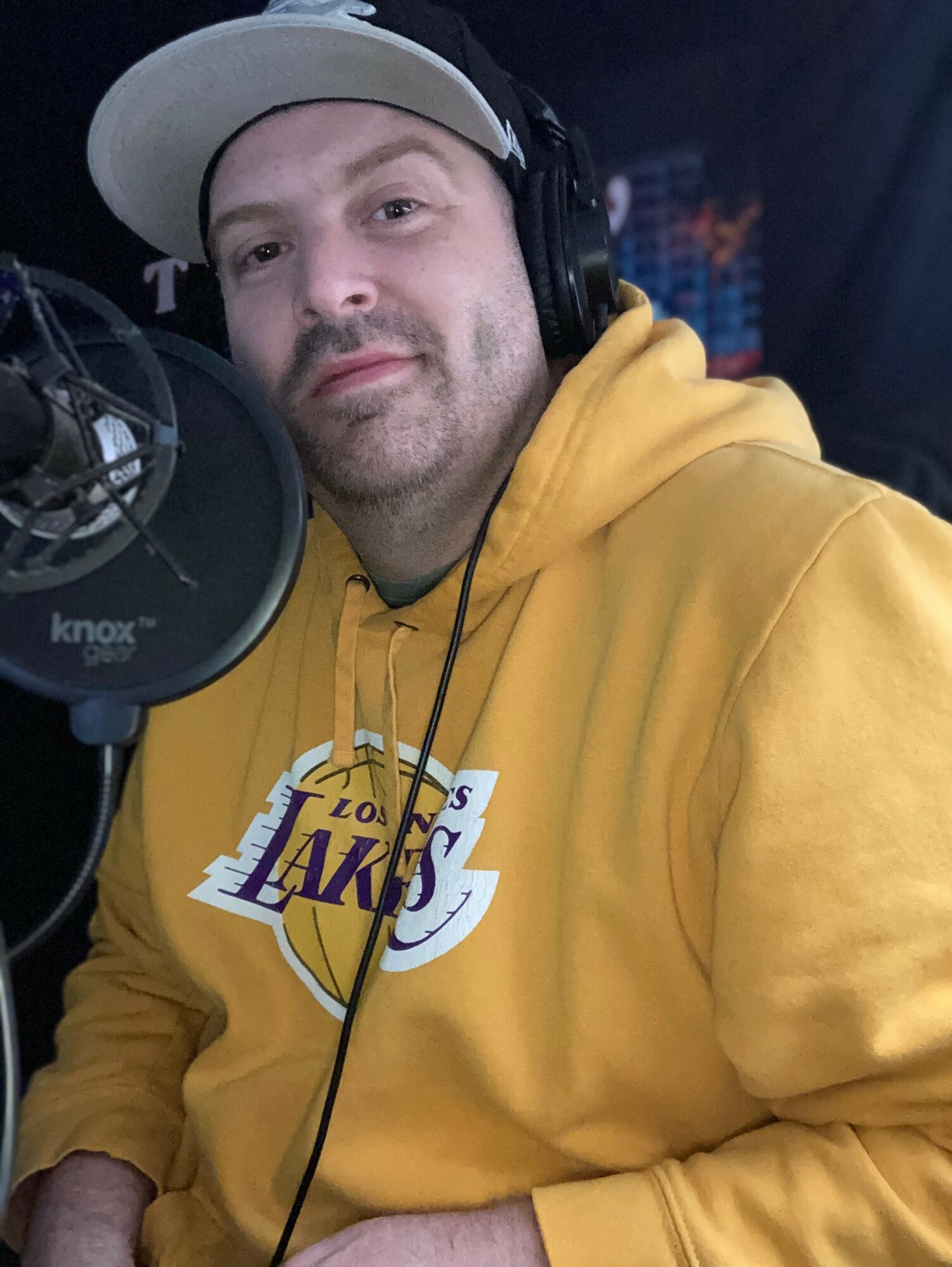 Man wearing a yellow Los Angeles Lakers hoodie and a white cap, speaking into a microphone in a recording studio.
