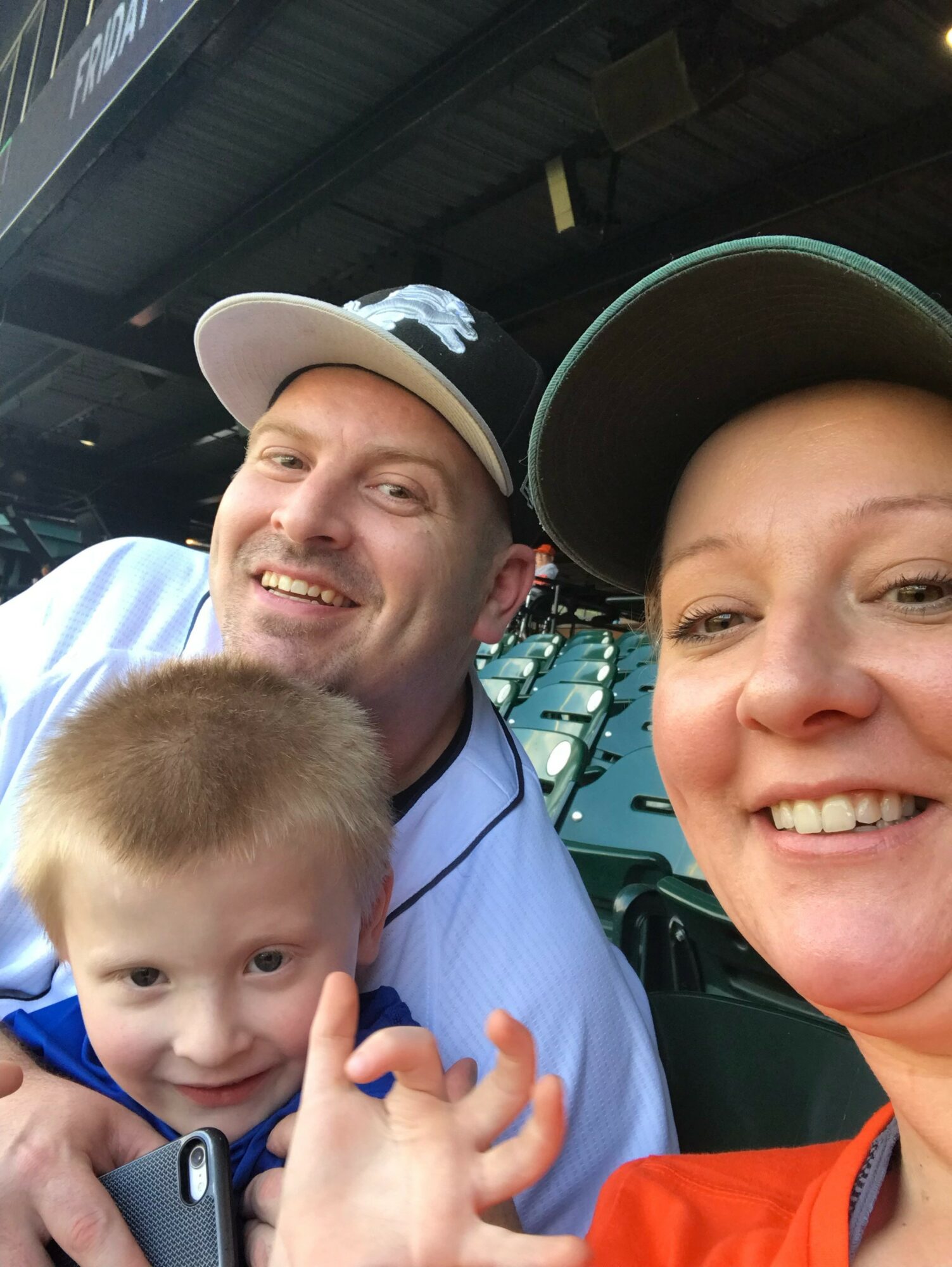 Three people smiling at a sports stadium, with a young child in the foreground, two adults behind, all wearing caps.