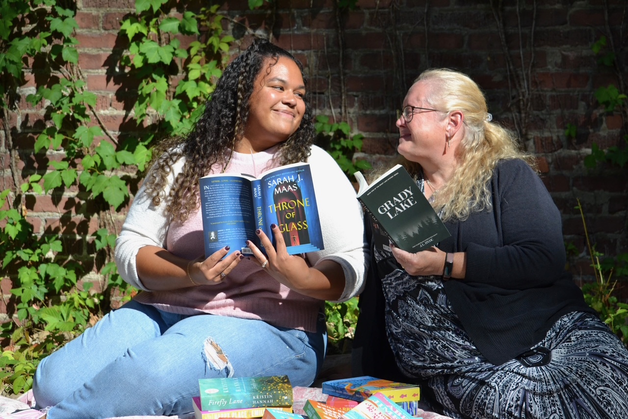 Two women sitting outdoors, smiling and holding books, with a brick wall and greenery in the background.