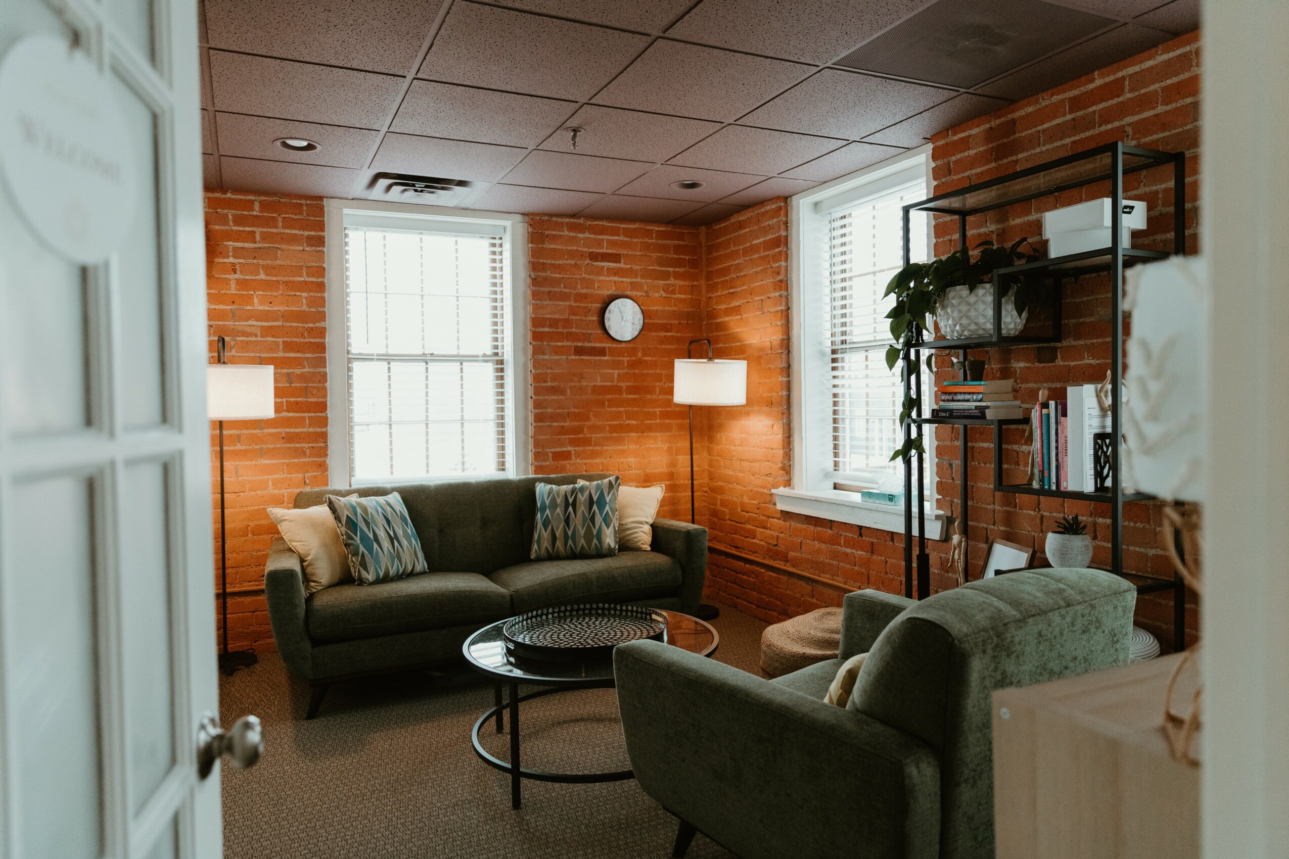Living room with brick walls, two windows, a sofa, armchair, round coffee table, and bookshelf, illuminated by natural and artificial light.