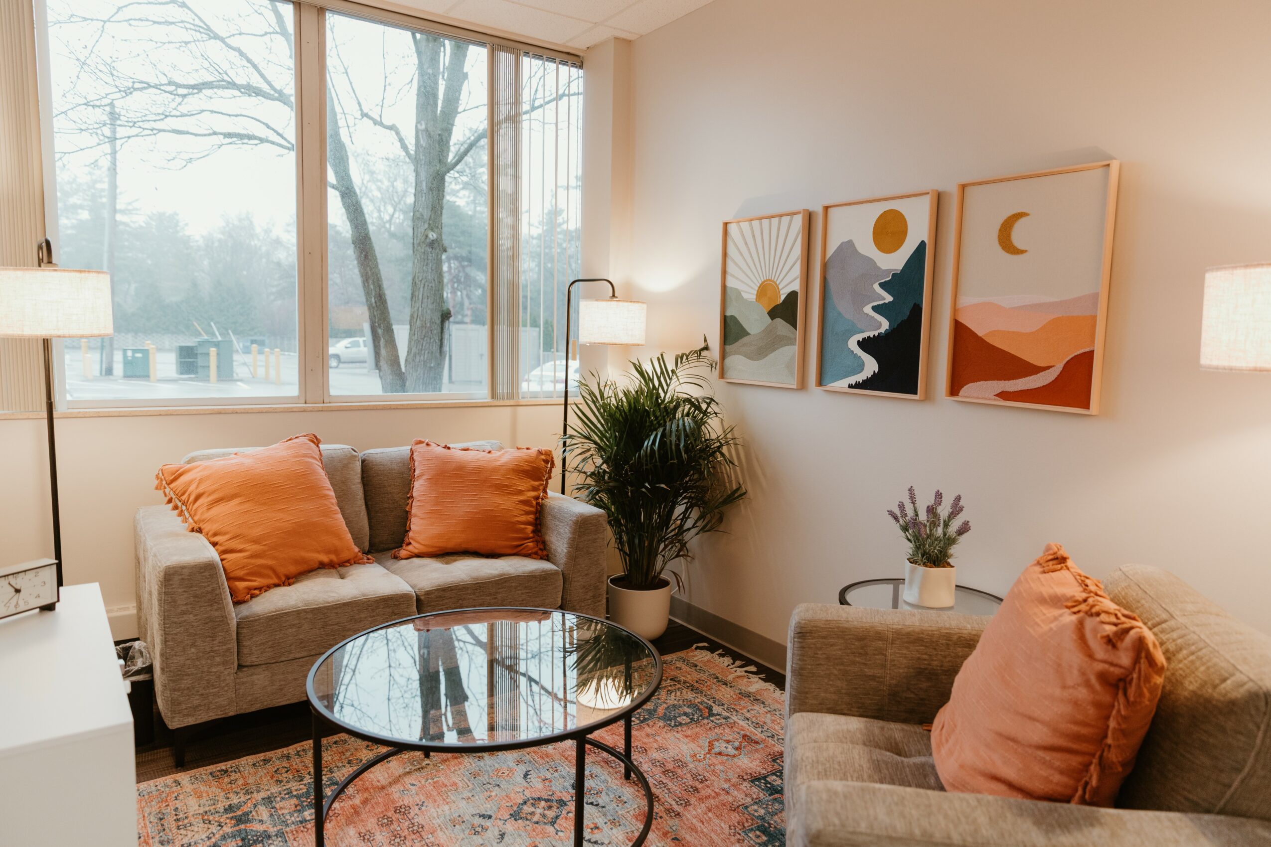 Living room with large window, two sofas with orange pillows, round glass coffee table, and framed artwork on wall.