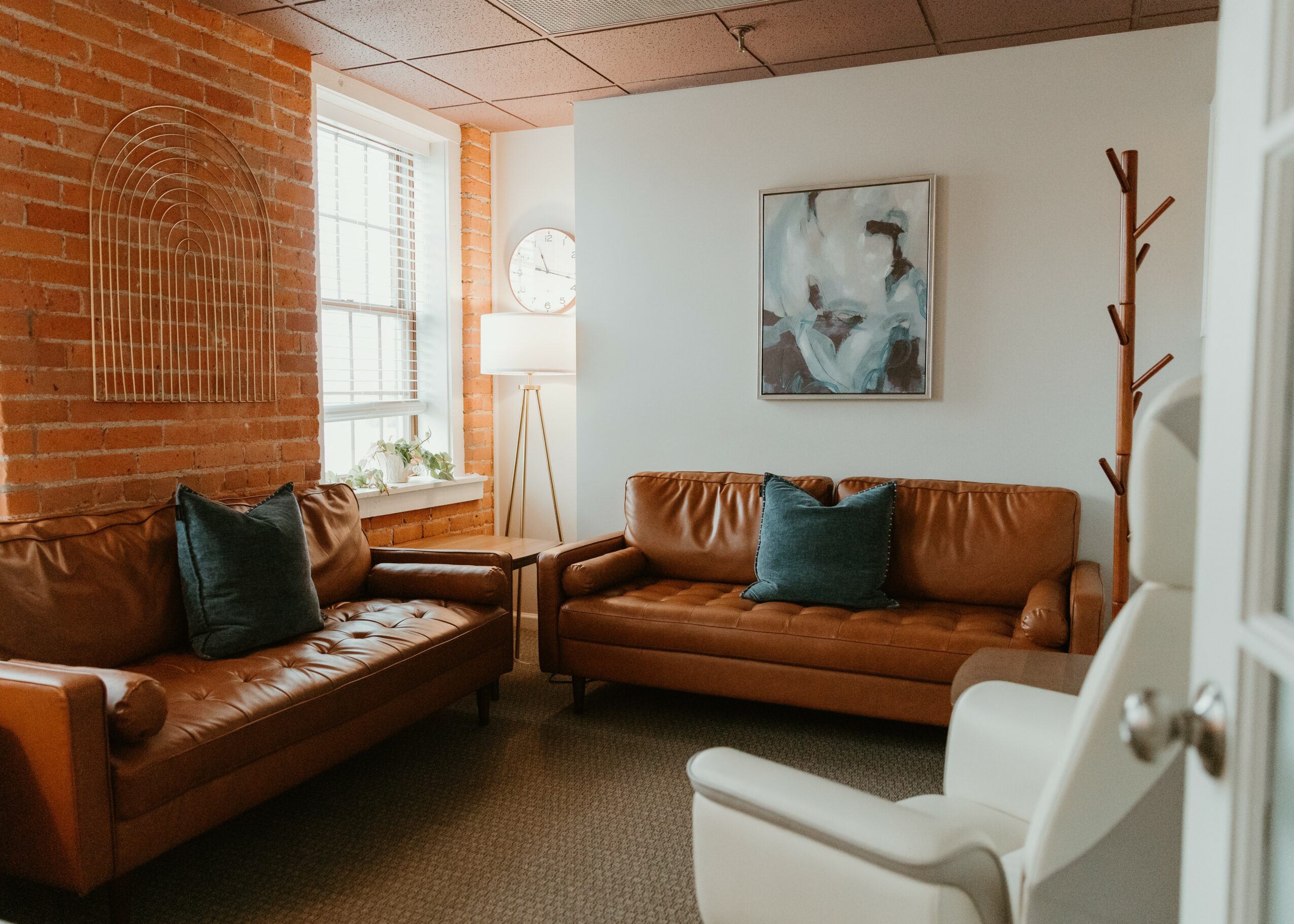 Living room with two leather sofas, a white armchair, a window, a lamp, a painting, and a coat rack.