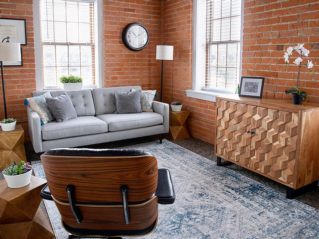 Living room with brick walls, large windows, gray sofa, wooden chair, patterned rug, and wooden sideboard.