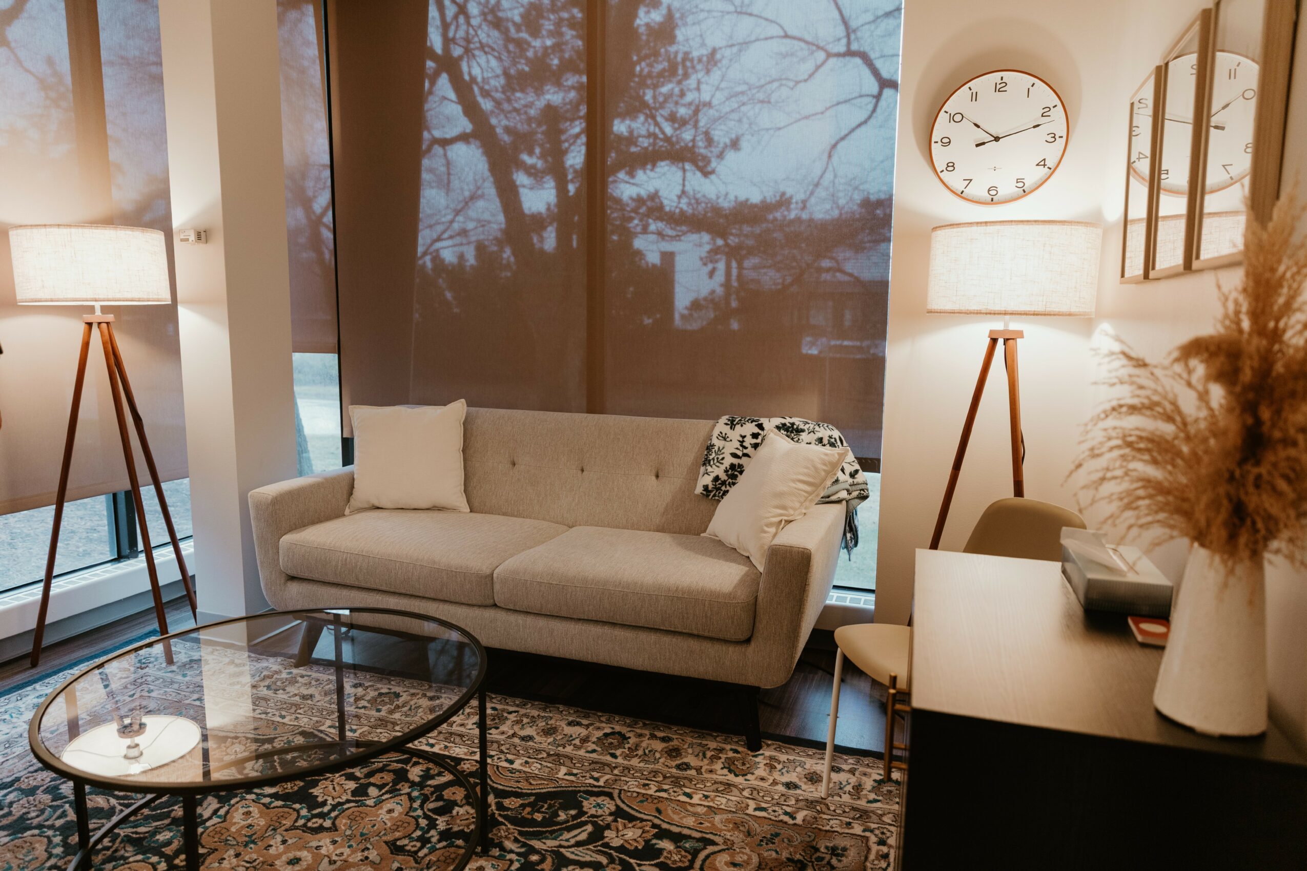 Living room with beige sofa, large window, two floor lamps, and a patterned rug, with a clock on the wall.