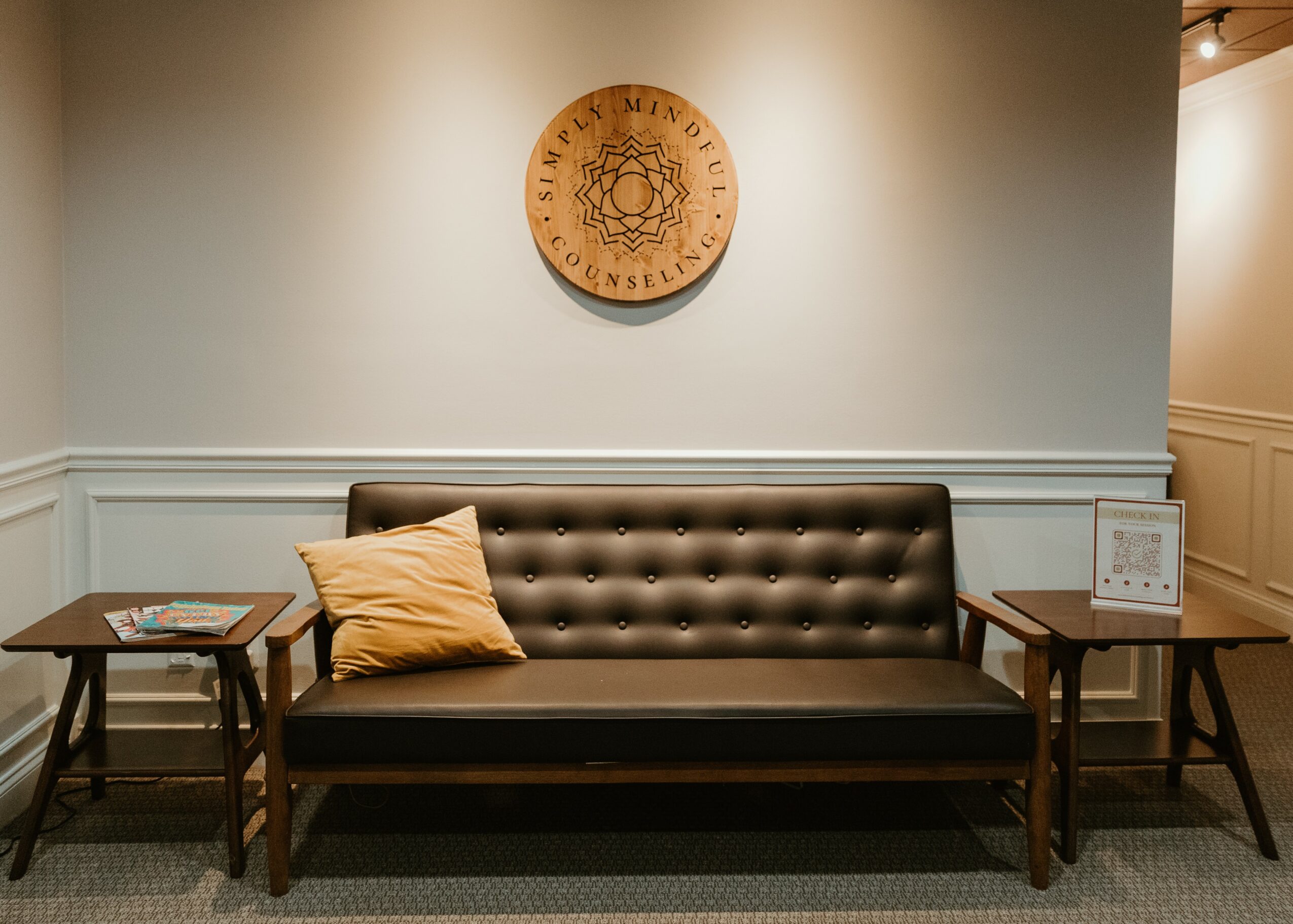 A black tufted sofa with a beige pillow, flanked by two wooden side tables, against a light-colored wall with a round wooden sign.