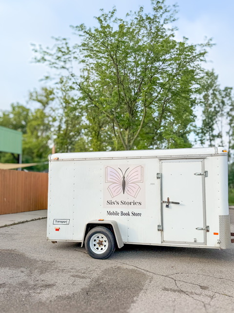 White mobile book store trailer parked on a paved surface with green trees in the background.